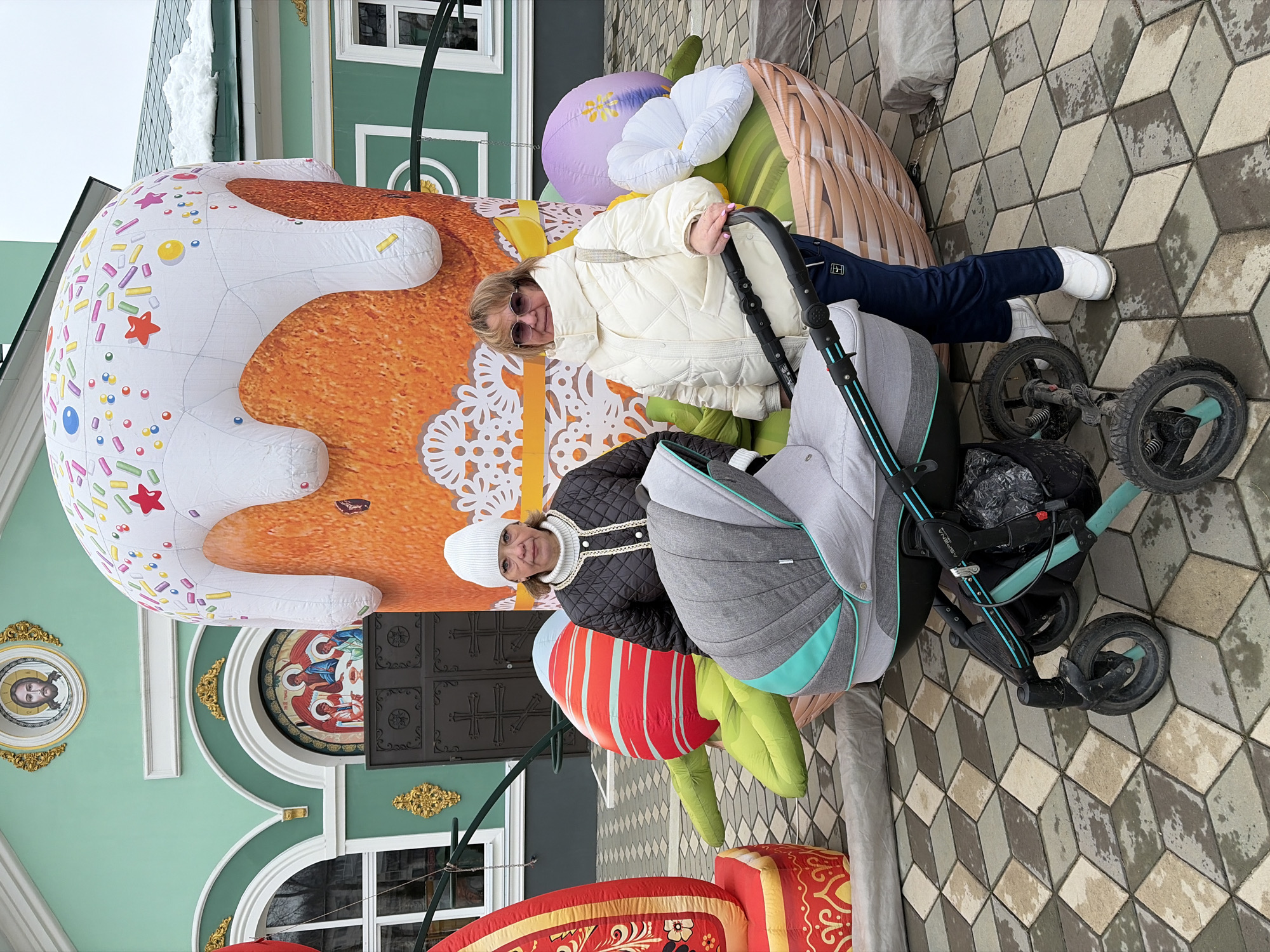 The image shows two women posing with a stroller next to an inflatable Easter display in Chaikovsky, Russia. The display includes a large inflatable Easter cake (kulich) decorated with colorful sprinkles and icing, inflatable flowers, and what appears to be an inflatable Easter egg.
One woman is wearing a white quilted jacket, dark pants, and white sneakers. The other woman is wearing a dark quilted jacket, a white hat, and a white turtleneck. They are standing/sitting next to a gray stroller with a teal accent.
The display is positioned in front of a building with a green facade, white trim, and a decorative window. The ground is paved with patterned gray and brown stones.