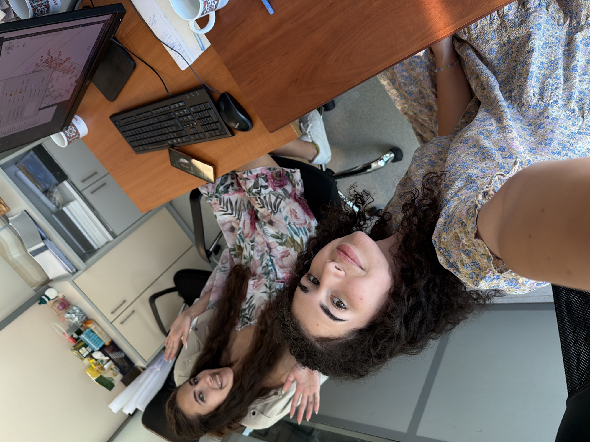 The image depicts two young women in an office setting, possibly in Sofia, Bulgaria. The woman in the foreground, with curly dark hair, is wearing a floral print dress and looking towards the camera with a slight smile. Her arm is extended, and a faint bracelet is visible. She is seated in a black office chair. Behind her, the second woman, with long dark hair, is also wearing a floral print dress and is seated in a similar office chair, looking towards the camera and waving. She is wearing a light-colored jacket over her dress.
The environment includes office furniture such as desks, chairs, and cabinets. On a desk in the background, a computer monitor displays a complex diagram, a keyboard, a mouse, and a smartphone are visible. Mugs are also present on the desk. In the background, cabinets with various items, including what appear to be office supplies and possibly snacks in baskets, are organized on shelves. The overall impression is of a casual office environment.