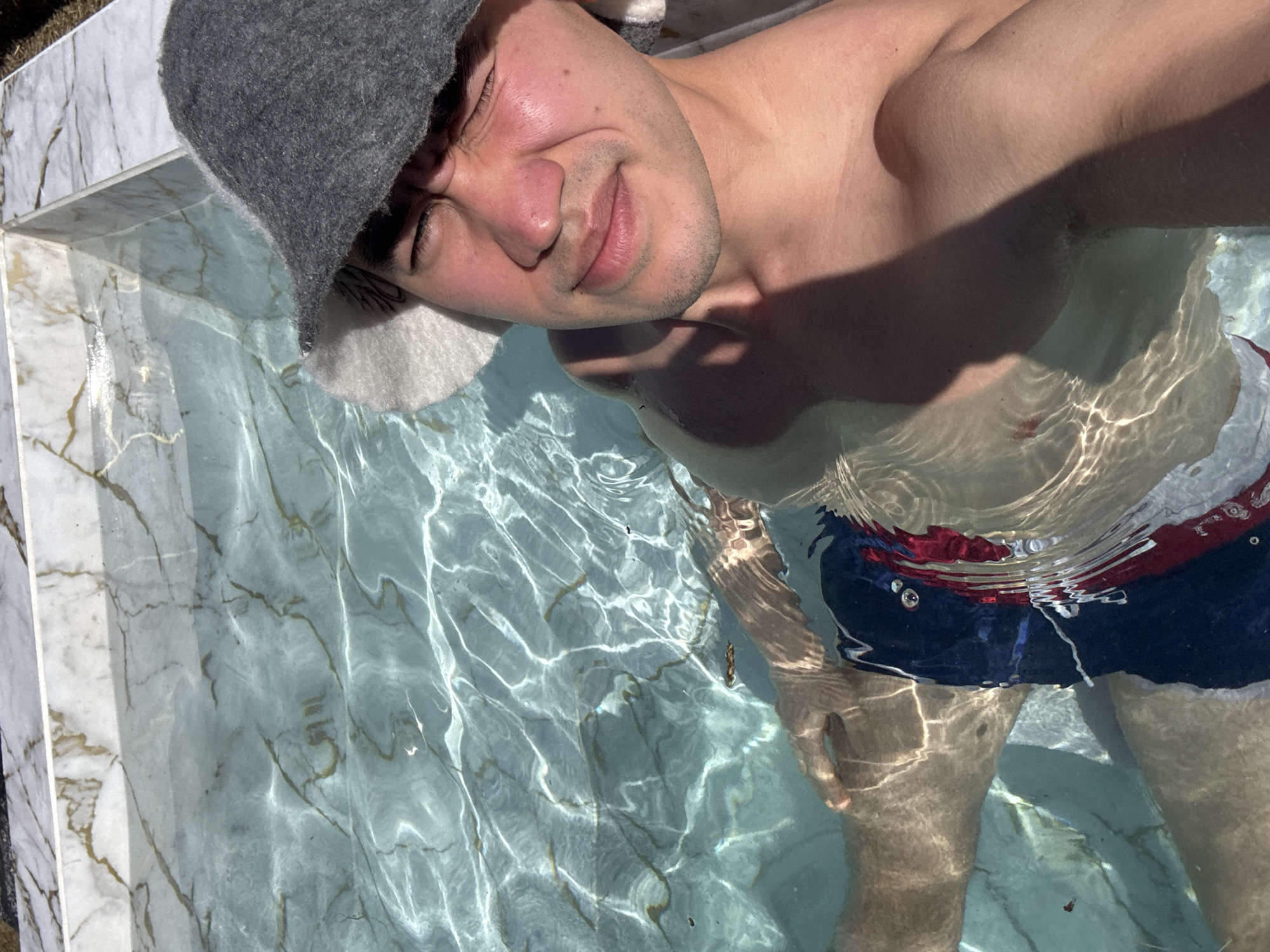 A man wearing a grey felt sauna hat and dark-colored swim trunks is partially submerged in a small, rectangular, marble-tiled pool. The water in the pool is clear and rippled, reflecting sunlight. The man is looking at the camera with a slight smile. The image was taken in Kazan, Russia.