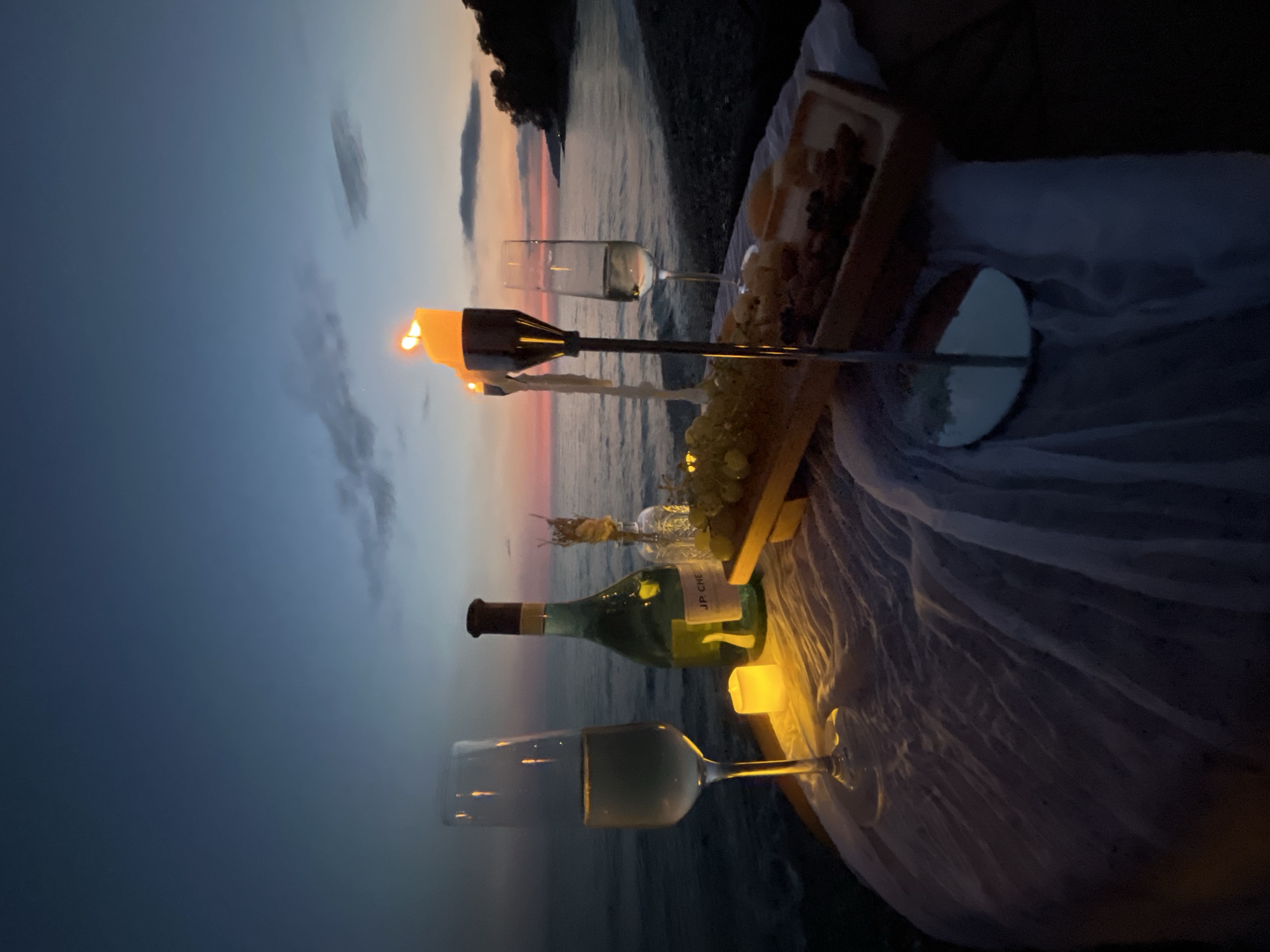 A romantic picnic setup is arranged on a table covered with white fabric near the water's edge. Two flutes of champagne are present, along with a bottle of JP. CHENET wine and a small vase with dried reeds. A wooden platter holds grapes and cheese. Two candles are lit, casting a warm glow. The scene is set against a dusky sky with hues of orange and purple, over a body of water. In the distance, a tree-lined shore is visible. The location is the city of Sochi, Russia, specifically a coastal area. There are no people visible in the image.