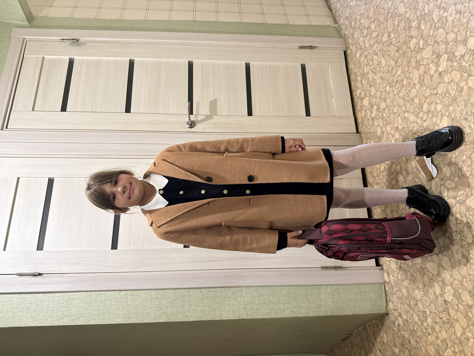 A young girl stands in a hallway, holding a backpack. She is wearing a brown coat over a dark dress with a white collar, white polka dot tights, and black patent leather shoes. The hallway features light wood doors with dark horizontal accents, and light green textured wallpaper on the left wall. The floor is covered with a light brown, pebble-like tile. The background includes a light fixture on the wall to the right. The scene is indoors and appears to be within a residential building. The girl is facing forward and looking at the camera. The image was likely taken in the city of Rusanovo, Russia.