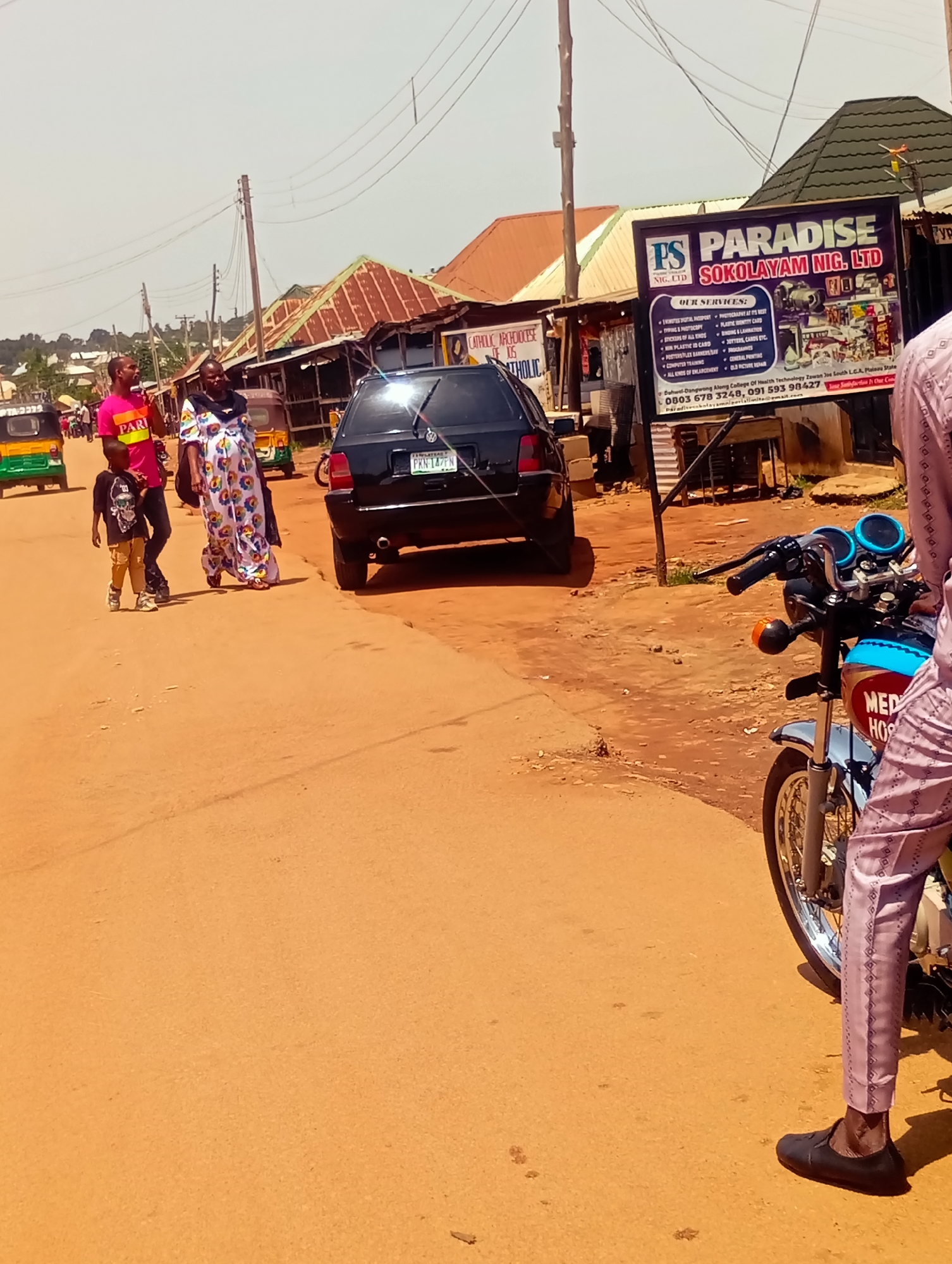 The image depicts a street scene in Du, Nigeria. In the foreground, a person wearing a patterned pink and purple outfit and dark shoes is partially visible, standing next to a blue and silver motorcycle. The motorcycle has a "MED HOS" inscription on its side. Further down the street, a black car is parked. To the left, three people are walking: a man in a pink shirt, a child in a black shirt and brown pants, and a woman in a colorful patterned dress. Behind them, there are several small structures with corrugated metal roofs and a yellow and green auto-rickshaw. A large billboard with the text "PS PARADISE SOKOLAYAM NIG. LTD" is visible on the right side of the street, displaying various services offered by the business, including passport photography, typing, stickers, and computer training. The ground is covered in reddish-brown dirt. Power lines stretch across the sky. The overall impression is of a busy commercial area on a sunny day.