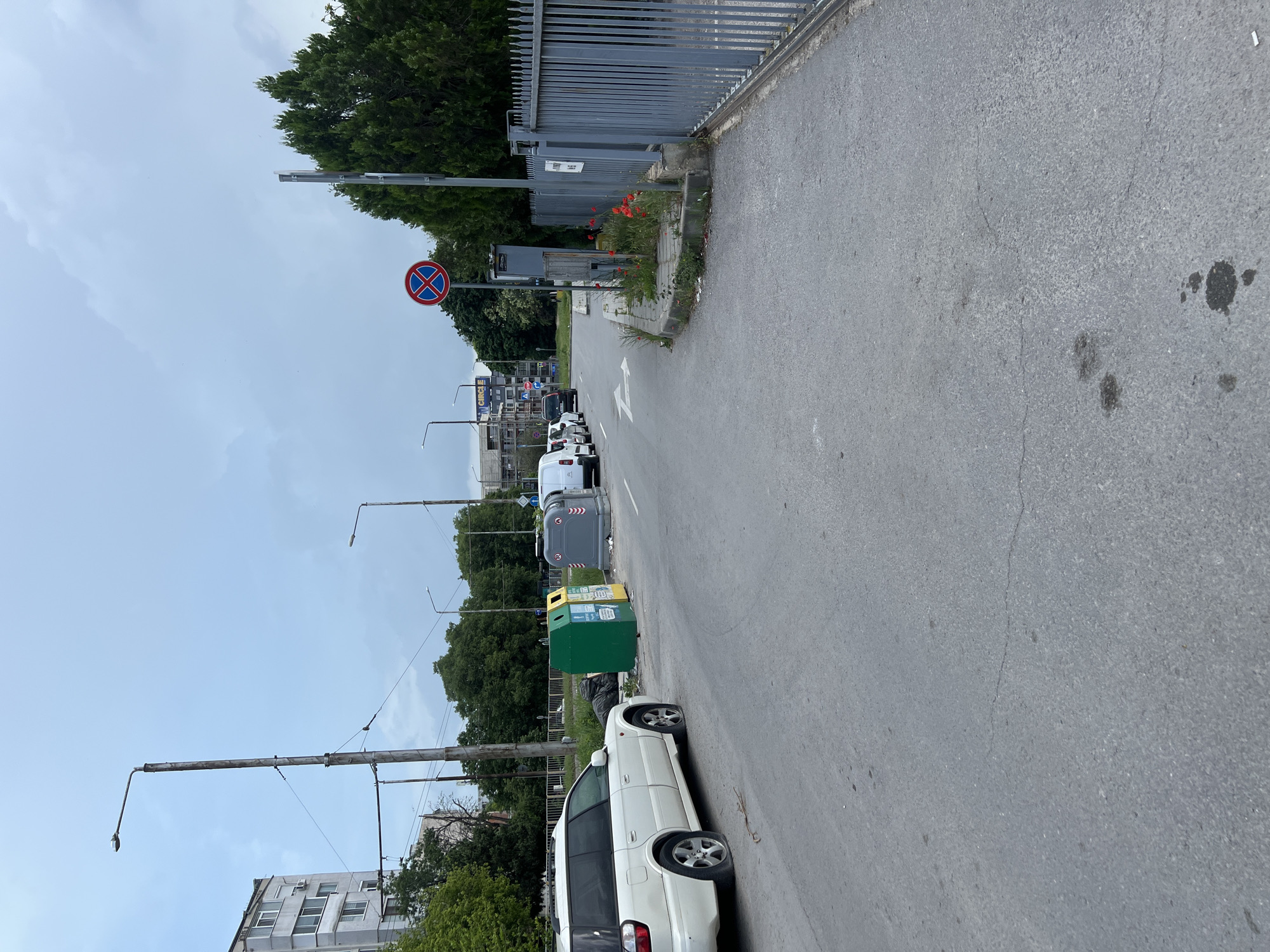 The image depicts a street scene in Varna, Bulgaria. The street has a grey asphalt surface with some visible stains and markings. A white minivan is parked on the right side of the road, partially obstructing the lane. Further down the road, several refuse bins of various colors (green, grey) are placed along the side of the road. A row of cars is parked further up the street. A "no stopping" sign is visible on the left side of the street, mounted on a pole. A grey metal fence runs along the right side of the street, adjacent to some greenery and red flowers. Street lamps line the left side of the road. The sky is overcast. A building is visible in the background. A gas station "CIRLCLE" is also visible on the left.