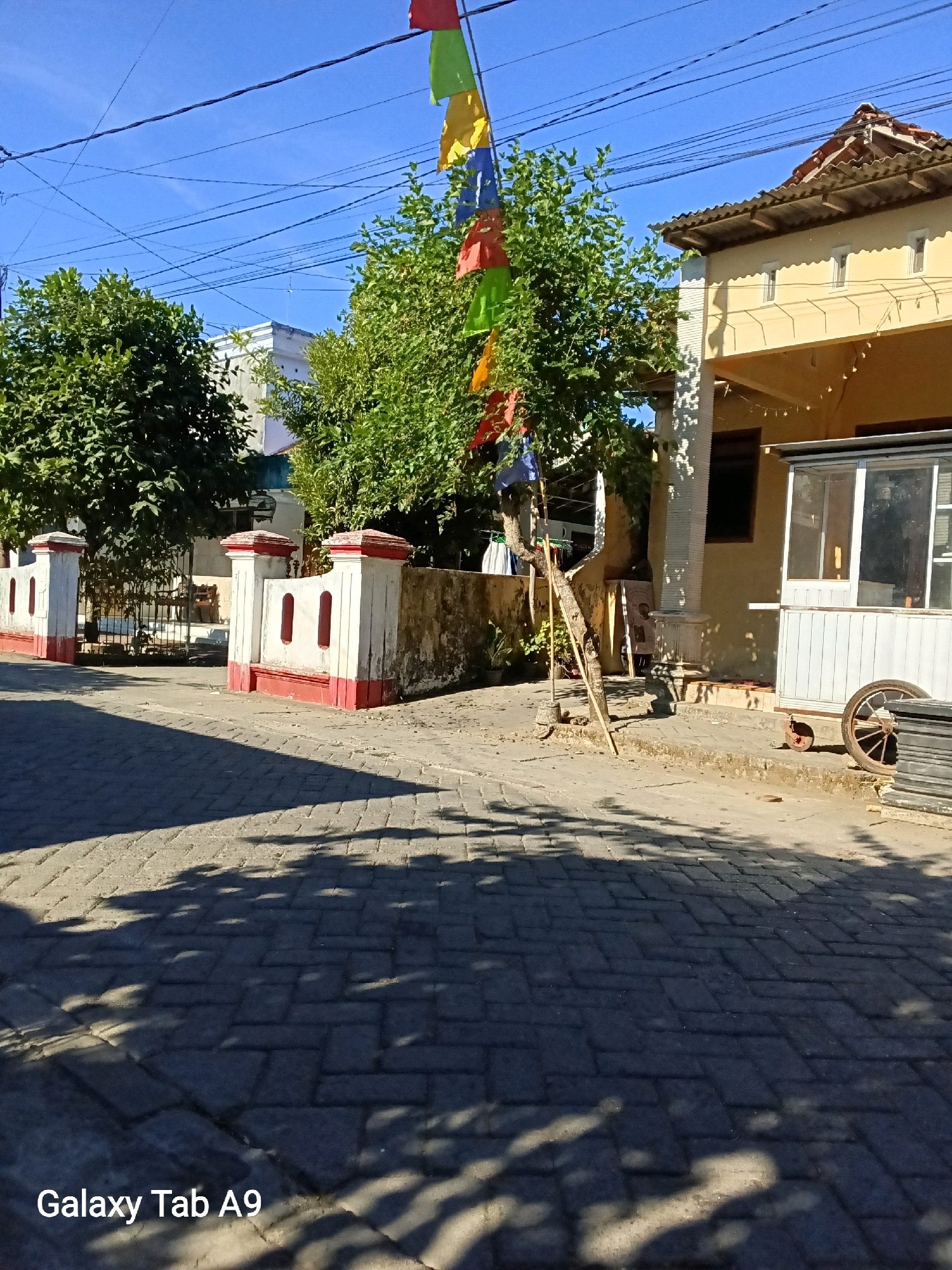 The image depicts a street scene in Kelutan, Indonesia. In the foreground, a brick-paved road is visible, with shadows of tree leaves cast upon it. To the left, a white and red-painted fence with decorative openings marks the entrance to a property. Beyond the fence, a large green tree stands, and in the background, a white building with several windows can be seen. To the right of the road, a yellow building with a tiled roof is partially visible. Adjacent to this building is a small white structure with a glass-paneled window. A large spoked wheel and stacked black objects are also present on the right side of the image. A string of colorful triangular flags is suspended from a pole and branches, adding a festive element to the scene. The sky is a clear blue, indicating a sunny day.