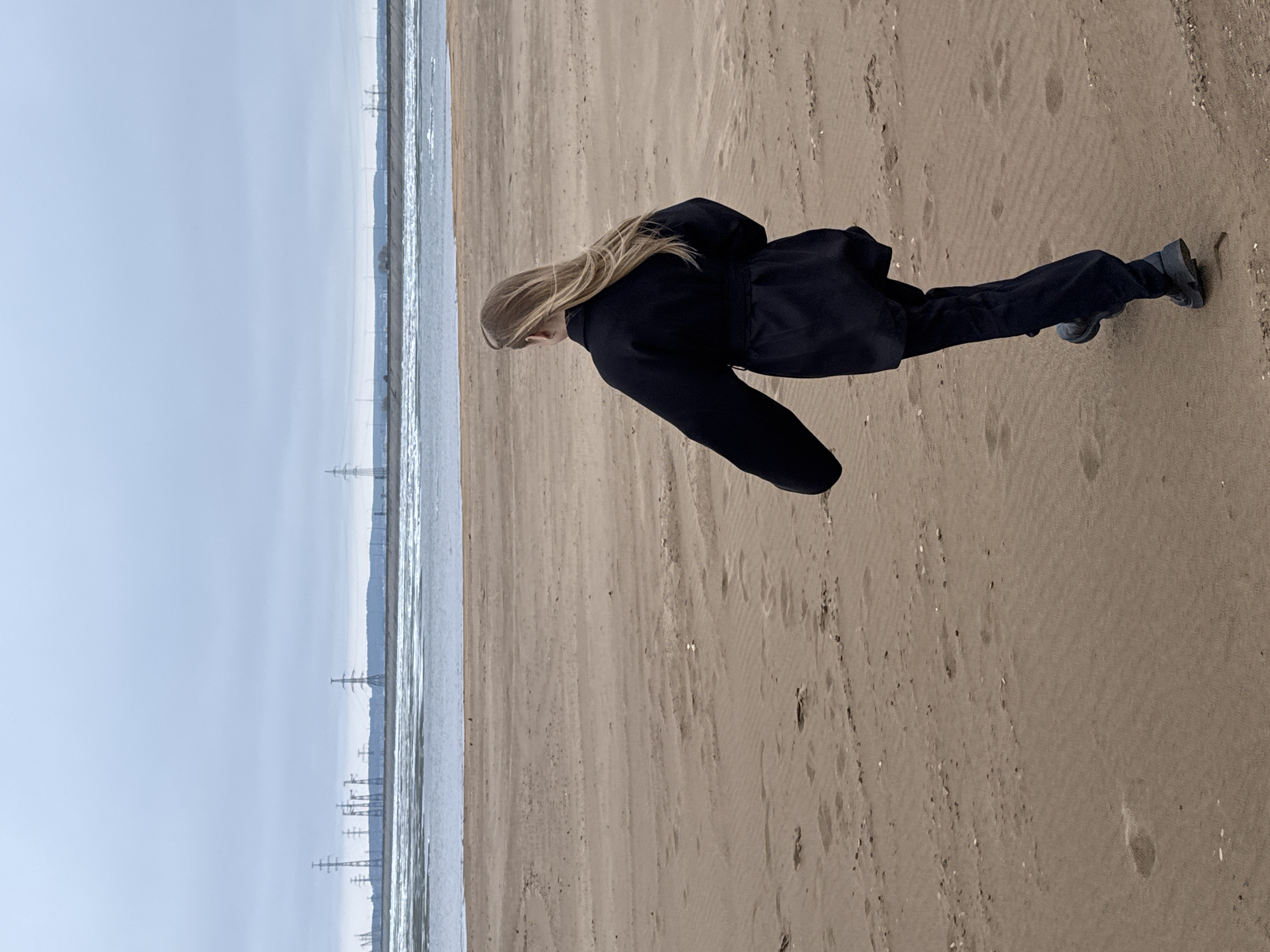 An image captures a person walking on a sandy beach next to a body of water under an overcast sky. The subject is a female with long, light-colored hair, seen from behind as she walks towards the water. She is dressed in a dark, possibly black, long-sleeved jacket or coat, dark trousers, and dark shoes or boots, suggesting cold weather.
The foreground is dominated by light brown sand, textured with ripples and numerous footprints. To the left of the sand, a strip of water appears partially frozen or icy, with lighter white sections indicating ice and darker patches showing open water. In the distance, across the expanse of water, a faint, low-lying landmass or coastline is visible. Several tall power transmission towers dot this distant horizon, suggesting a nearby industrial or developed area. The sky is a uniform, pale grey, indicating an overcast daytime. There are no other discernible activities or events taking place, and no visible text is present. The scene conveys a cool, possibly chilly, coastal environment, consistent with the location context of Noviy, Russia.