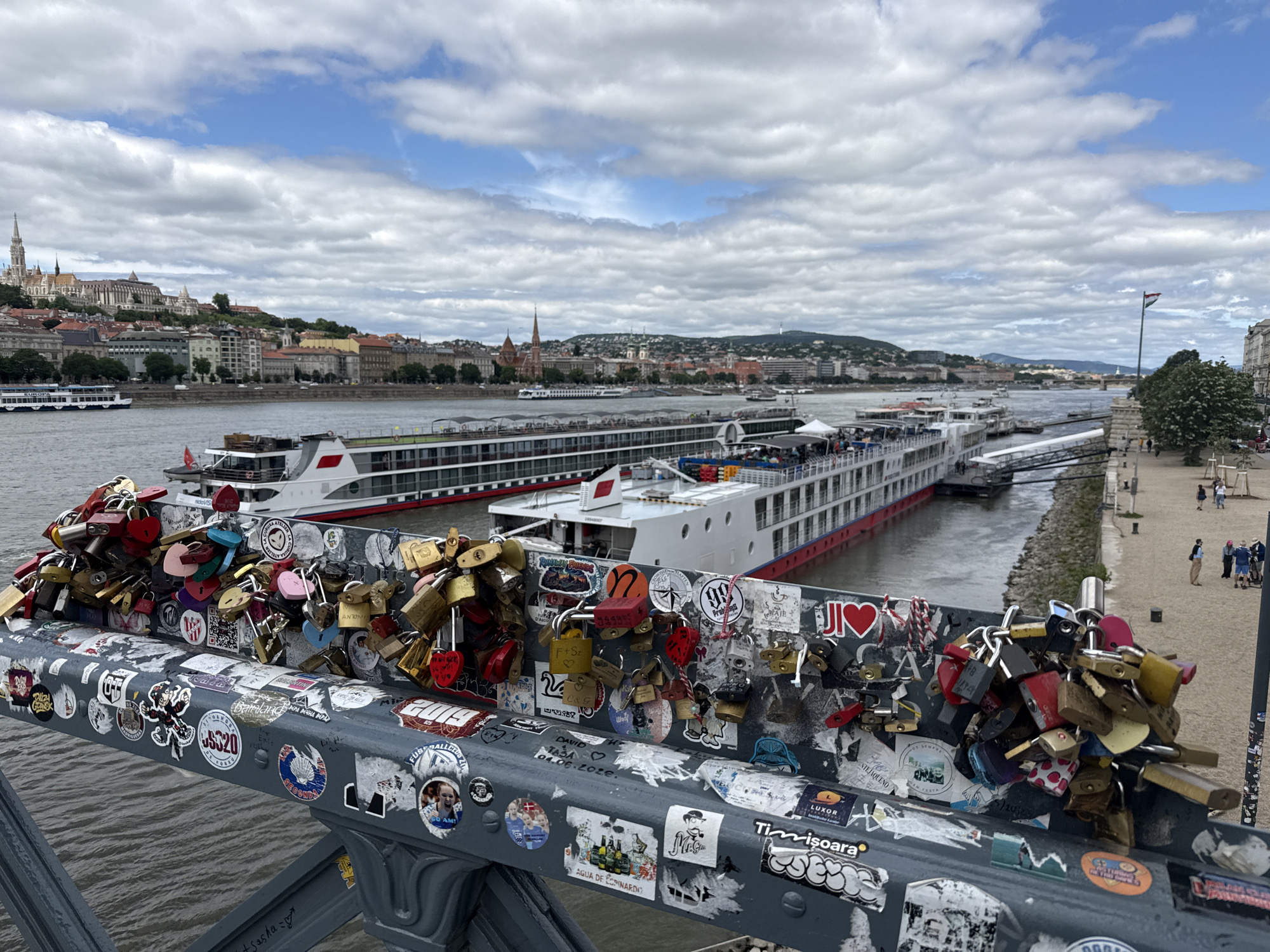 The image shows a close-up view of a railing adorned with numerous padlocks, many of which are colored and some are heart-shaped. Various stickers are also affixed to the railing. In the background, a wide river flows, with several large cruise ships docked along its banks. Across the river, a cityscape with numerous buildings and spires is visible, suggesting an urban setting. The sky is partly cloudy. The foreground railing appears to be part of a bridge.