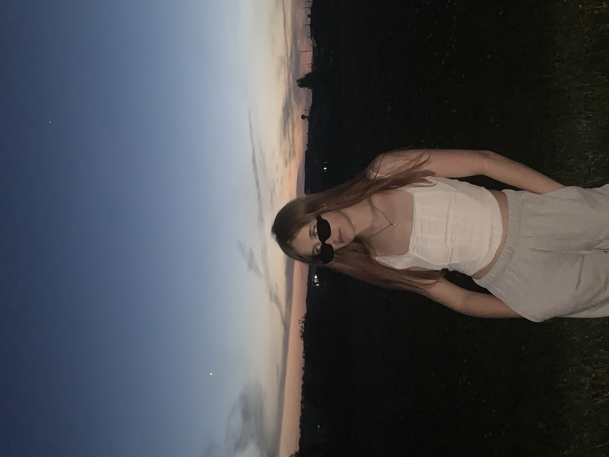 A sideways photograph depicts a person in sunglasses and a white top standing in what appears to be a grassy area at dusk. The person is oriented diagonally across the frame, with their head near the center and their lower body towards the right edge. The sky, visible on the left, transitions from a light orange near the horizon to a deep blue further up. The background on the right appears dark, possibly due to trees and vegetation. The image is rotated 90 degrees counterclockwise. The location, Фоки, Russia, is not discernible from the image itself.
