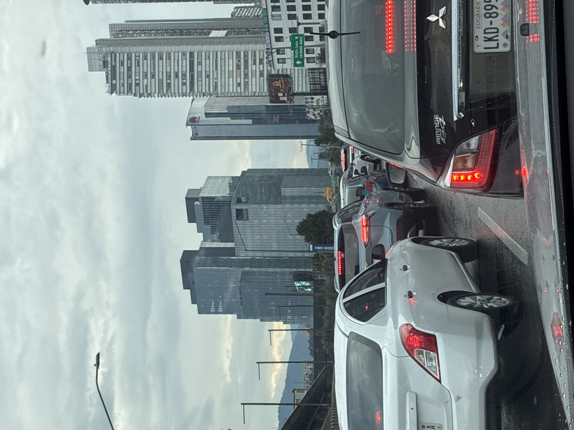 The image depicts a traffic jam on a roadway in Mexico City, Mexico. Several high-rise buildings, including one with a "Huawei" logo, can be seen in the background, indicating an urban area. A road sign pointing to "TOLUCA Cuota / SALVADOR AGRAZ" is visible. In the foreground, multiple cars are congested, with their tail lights illuminated. The black car immediately in front of the camera is identified as a Mitsubishi Mirage with license plate "LKD-895" from the State of Mexico. The weather appears overcast, and there are water droplets on the window, suggesting rain.