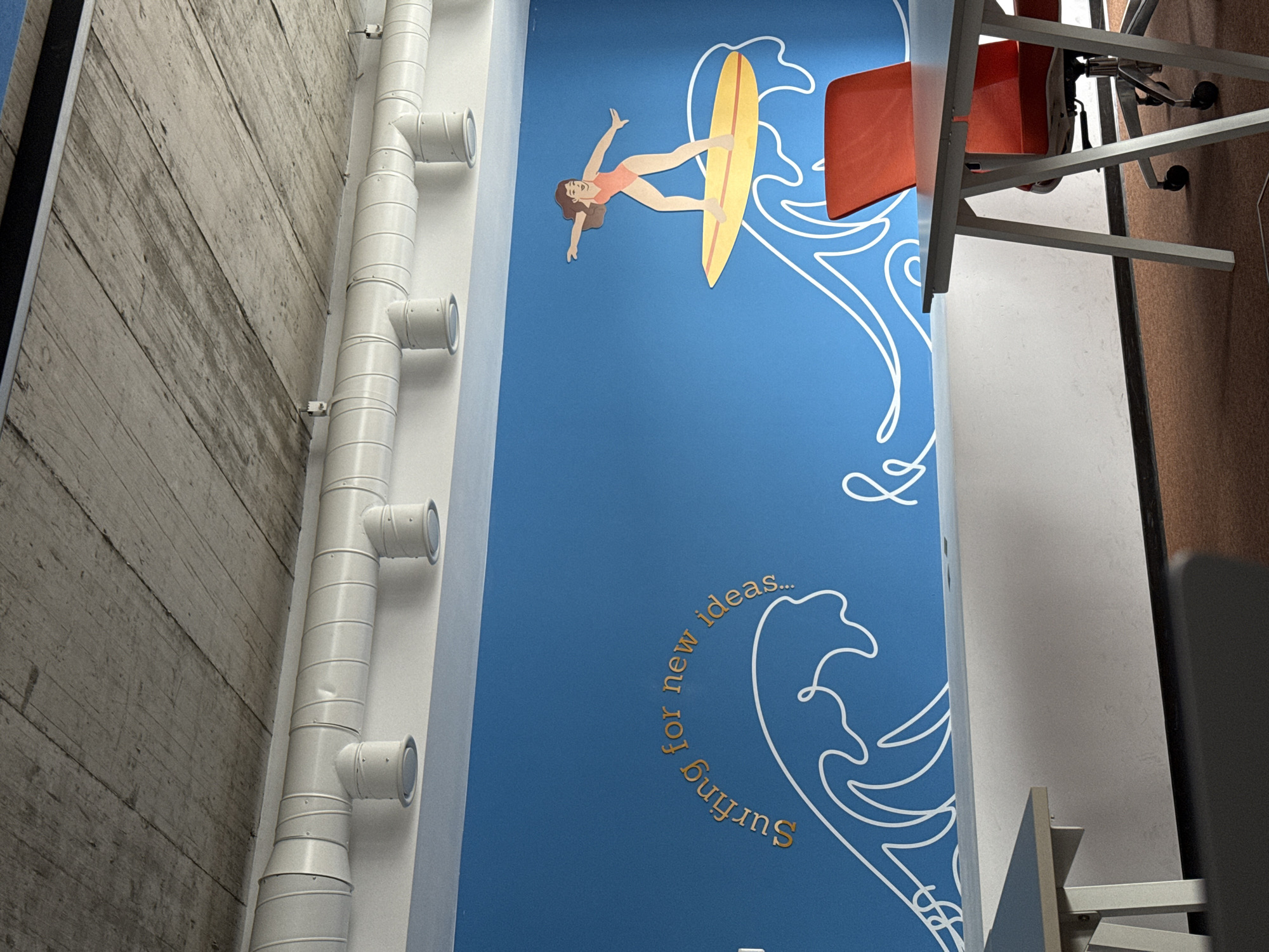The image shows a section of a wall in what appears to be an interior space. The wall is painted bright blue and features a cartoon-like illustration of a surfer riding a wave, along with the text "Surfing for new ideas..." rendered in a golden hue. Adjacent to the blue wall is a grey textured wall with a white pipe fixture running vertically. To the right of the blue wall, office furniture can be seen including a red office chair. The location appears to be the Innovation Starter Varna co-working space at 1 Tsar Boris III Blvd in Varna, Bulgaria.