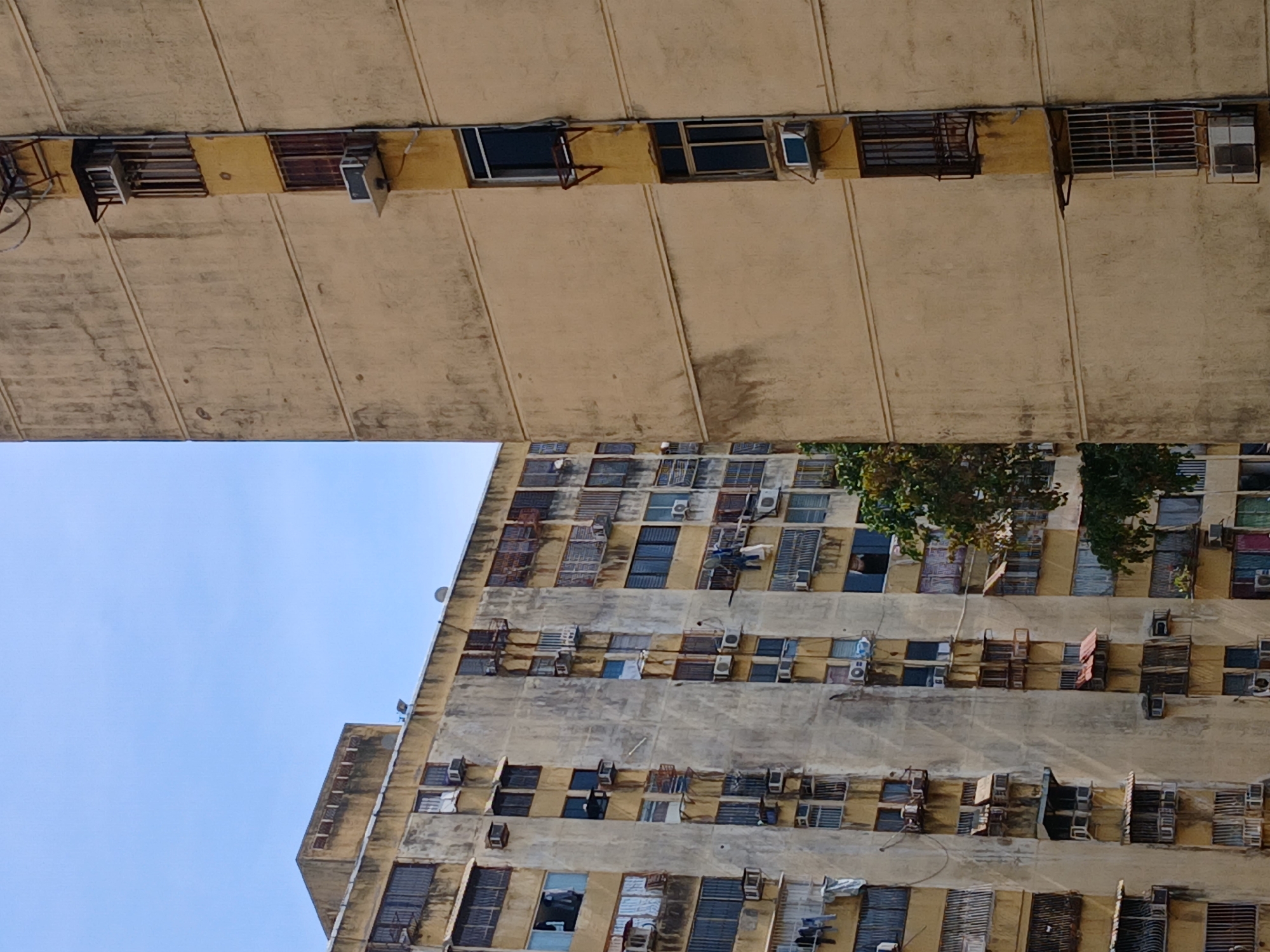 The image captures an upward-looking perspective of two multi-story residential buildings under a clear, bright blue sky, typical of an urban environment in Ciudad Guayana, Venezuela.
In the upper half of the frame, the underside of a large, cantilevered concrete structure, likely an extensive balcony or overhang of a nearby building, dominates. This surface is a weathered light beige or yellowish hue, marked by extensive staining, dirt, and visible construction lines, indicating age and exposure to elements. Along the vertical edge of this overhead structure, multiple windows are visible, some fitted with dark security bars or metal grilles, and various window-mounted air conditioning units are installed. Exposed wiring and metal brackets are also apparent.
Below this foreground structure, and against the clear sky, a tall, multi-story residential building rises. This building, also constructed from concrete, shares a similar weathered appearance and light beige-yellow color. Its facade is densely arrayed with numerous windows, most of which are protected by security bars or grates. A striking feature is the proliferation of window-mounted or wall-mounted air conditioning units, nearly one per window, underscoring the warm climate. Some windows are open, others closed, and faint suggestions of items or clothes are visible near a few openings. Patches of green tree foliage are visible clinging to the building's mid-section on the right.
The scene is set during bright daylight under a sunny sky. No individuals are visible, but the architectural details, and the sheer number of air conditioning units, are strong indicators of a densely populated urban residential area in a hot climate. No discernible text is visible in the image.