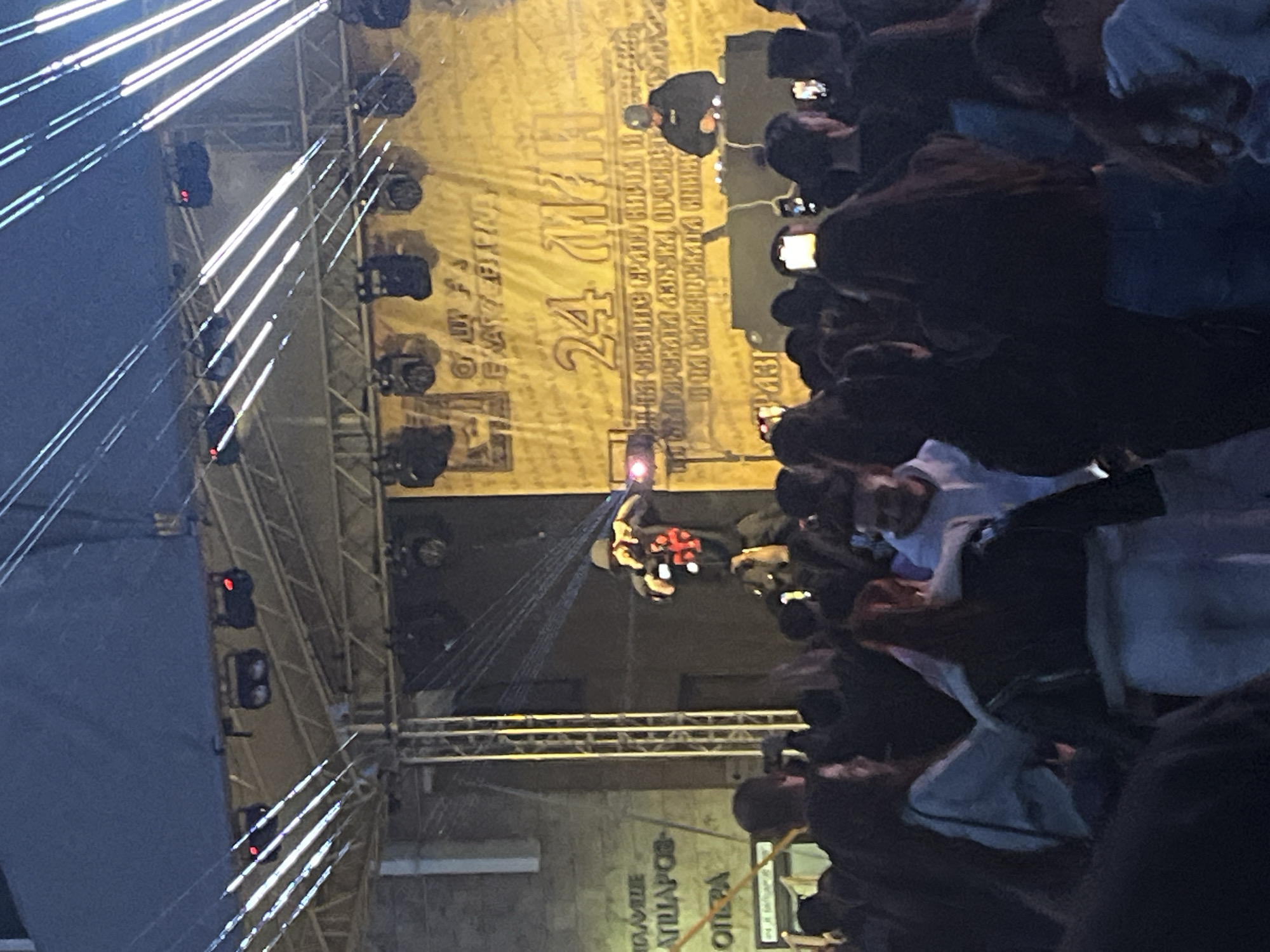 The image depicts an outdoor event in what appears to be Blagoevgrad, Bulgaria. A stage is set up with lighting rigs visible on the left. A screen behind the stage features text. People are gathered in front of the stage, some holding up their phones to record or take pictures. There are performers on the stage. The building with Cyrillic script on the bottom of the image is the theater building in Blagoevgrad.