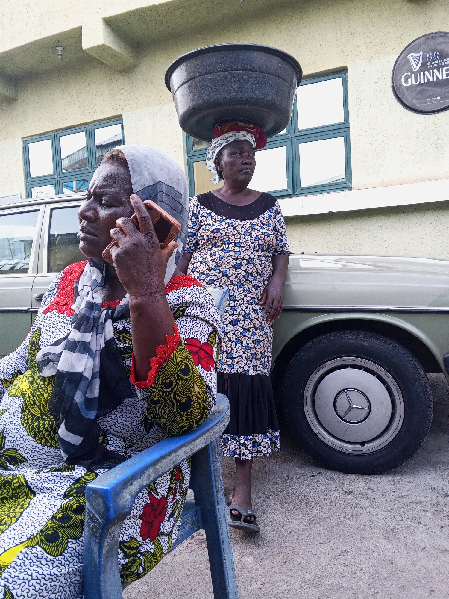 Two women are featured in an outdoor setting, likely in Zango-ndaji, Nigeria, against a backdrop of a light-colored building and a vintage car. The scene is illuminated by natural daylight.
In the foreground, a woman sits on a blue plastic chair, holding a rose-gold smartphone to her right ear, appearing to be in conversation. She wears a vividly patterned dress with yellow, black, and red motifs on a white background, featuring red lace detailing on the sleeves. A grey and black patterned headscarf covers her head. Her expression is serious and attentive, indicating focus on her call.
To her right and slightly behind, another woman stands beside a light green Mercedes-Benz sedan. She expertly balances a large, black basin on her head. This woman is dressed in a blue and white floral-patterned dress with a black bodice and a black ruffled hem, complemented by a colorful patterned headwrap. She looks towards the left, outside the frame. The front wheel of the Mercedes-Benz, identifiable by its hubcap bearing the brand's logo, is prominent.
The background includes a light yellow-green building with several dark-framed windows. A round, black Guinness sign is affixed to the building, clearly showing the text "ESTD 1759," "ST. JAMES'S GATE DUBLIN, IRELAND," and "GUINNESS." The ground is a paved concrete surface. A black sandal, possibly an Adidas brand, is visible on the foot of the standing woman.
