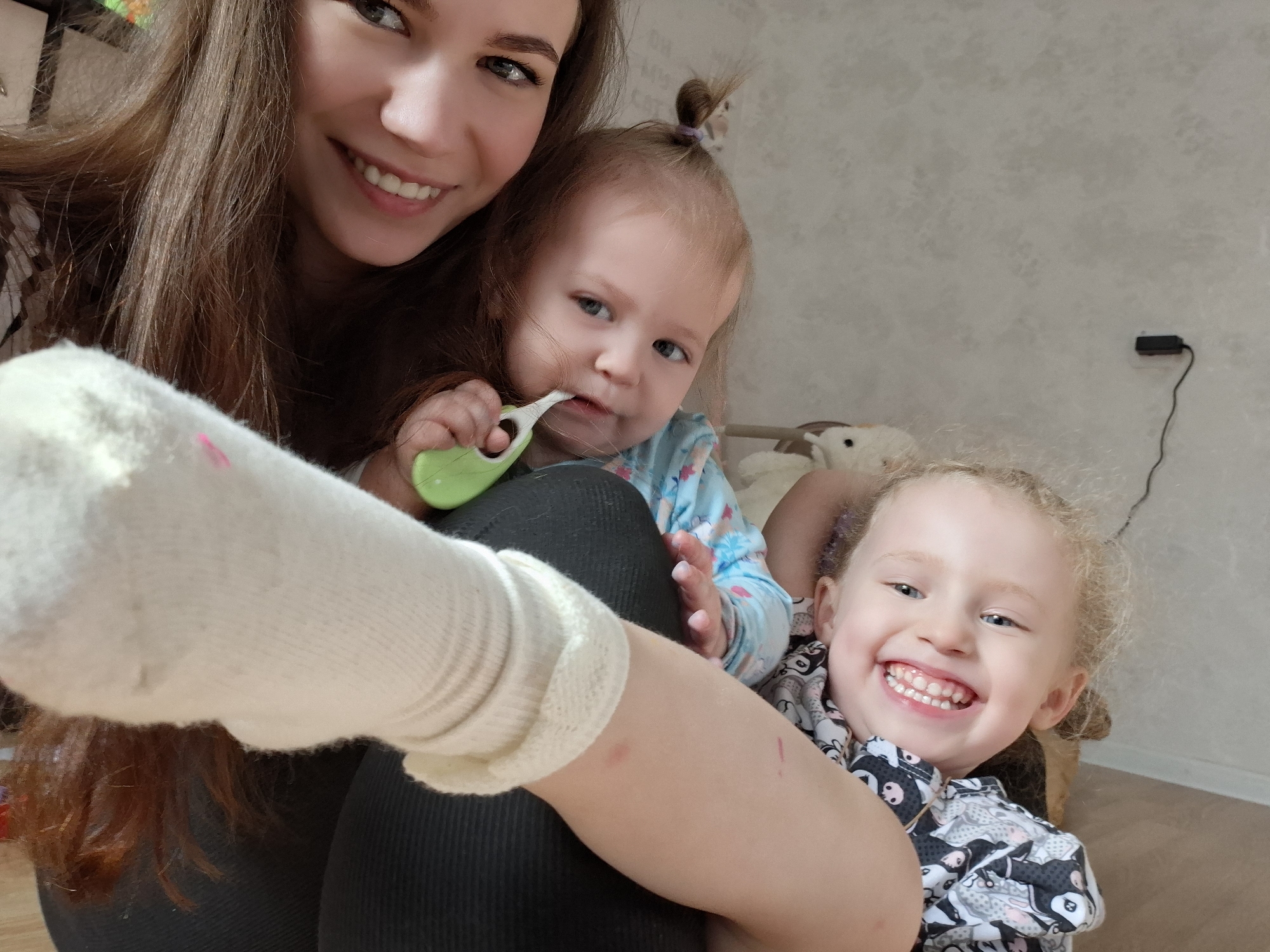 A woman and two young children pose for a selfie. The woman, with long brown hair, smiles. A baby with blonde hair in a ponytail nibbles on a green toothbrush. A toddler with curly blonde hair smiles brightly, revealing their teeth. The setting is a room with pale gray walls. A white sock is prominently displayed in the foreground. The location is Chaikovsky, Russia.
