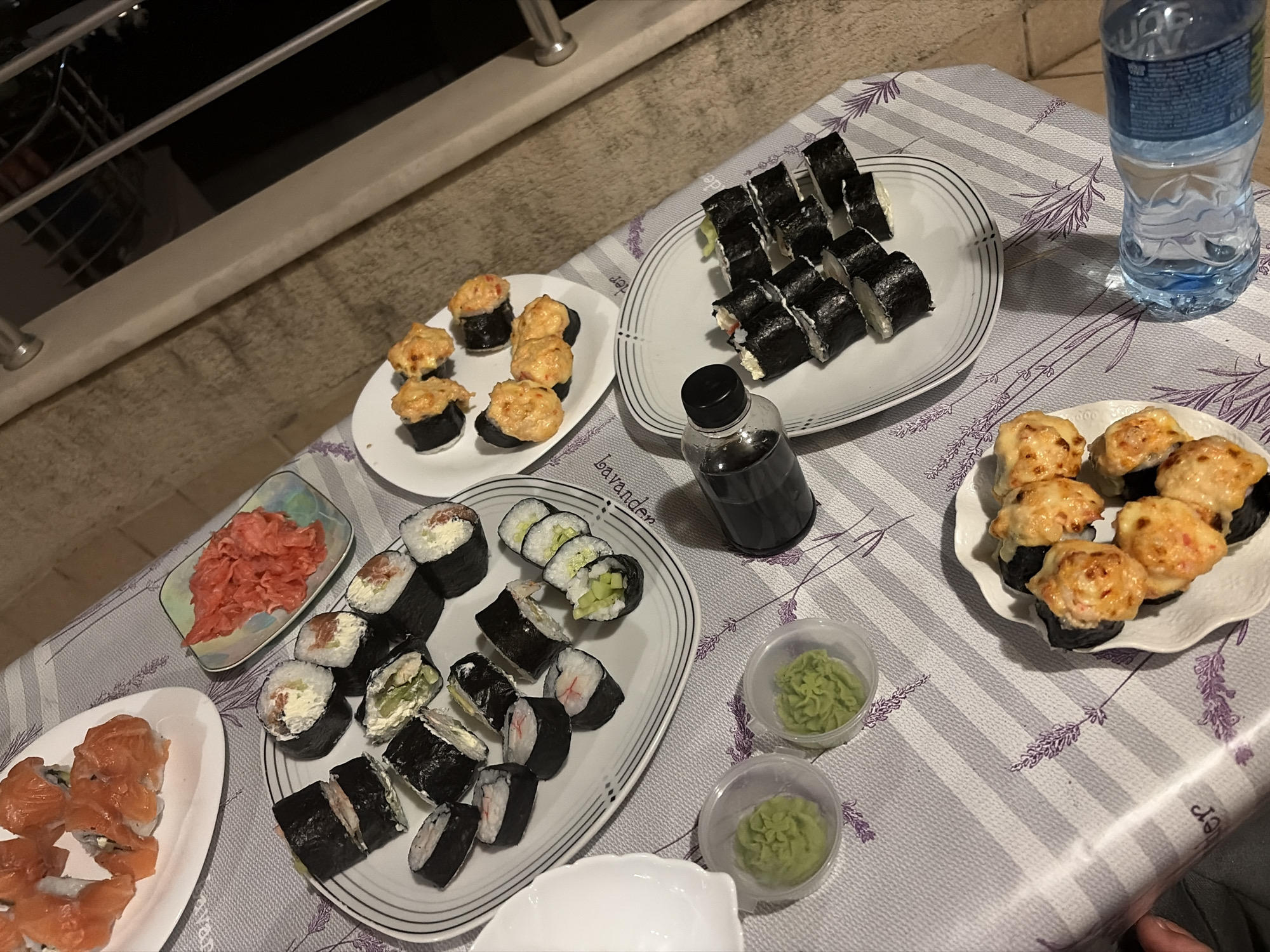 A table is set with a variety of sushi dishes and condiments, suggesting a meal is in progress or about to begin. The arrangement indicates a casual dining setting, possibly outdoors on a balcony or terrace, given the lighting and background elements that hint at a view of buildings and railings typical of urban environments, possibly in Budva, Montenegro.
The table is covered with a lavender-patterned tablecloth. Several plates of sushi are visible. One large white plate contains several pieces of sushi rolls with visible salmon and cream cheese. Another white plate holds various sushi rolls, some with dark nori wrappers and others with rice on the outside, with fillings like cucumber and imitation crab. A smaller rectangular dish holds pink pickled ginger. Two small plastic containers with green wasabi are positioned near these plates.
Further along the table, a white plate displays several pieces of baked sushi rolls, topped with a golden-brown, creamy sauce. To the right, a bottle of water is placed on the table. A dark bottle of soy sauce is also present. The overall scene is well-lit, possibly by artificial light given the contrast in the background, but the overall impression is that of an enjoyable meal shared in a pleasant setting. The image appears to have been taken from a slightly elevated, angled perspective.