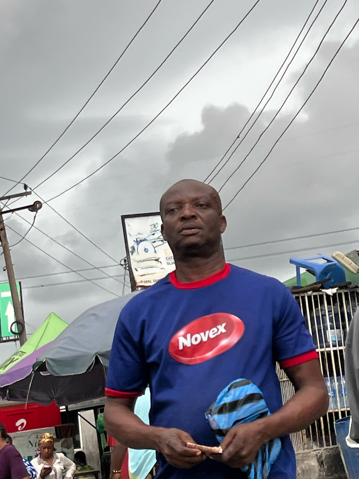 An outdoor street scene in Ado, Nigeria, features a dark-skinned man in the foreground wearing a blue t-shirt with a red collar and a red oval logo that reads "Novex." He is holding a folded blue and black striped bag in his right hand and appears to be holding a small object in his left hand. The man's head is shaved, and he is looking slightly to his right. In the background, several utility lines are visible against a cloudy sky. To the left, a green and purple umbrella shades a stall. A sign with text and an image is mounted on a pole behind the man. A blue plastic chair and a metal fence with bottles are visible further in the background.