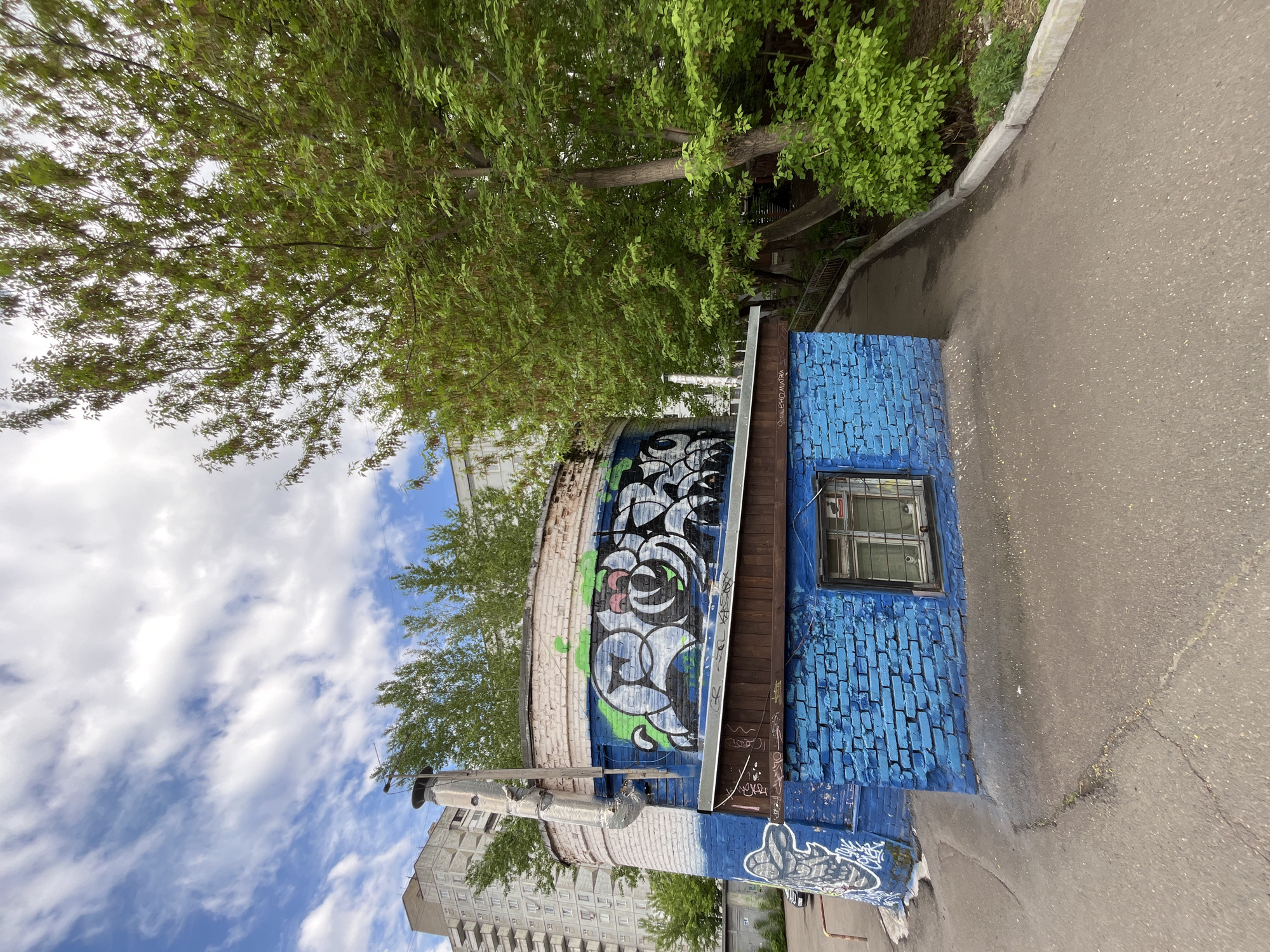 The image shows a blue brick building with graffiti, located in Krasnoyarsk, Russia. The building has a window with metal bars. The graffiti is mostly black, white, and blue, with some green accents. A cylindrical structure, also with graffiti, adjoins the blue building. A metal pipe runs up the side of the cylindrical structure. The building is situated next to a paved road. Trees and a section of a larger building are visible in the background. The sky is partly cloudy.