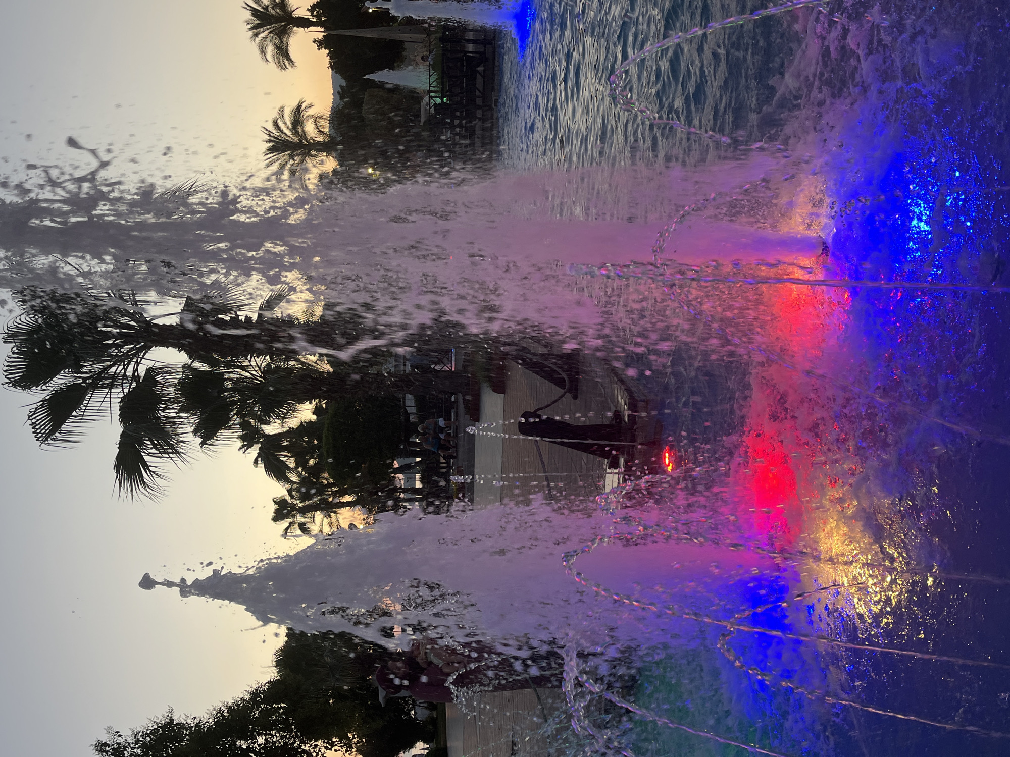 The image shows a water fountain lit with red, blue, and pink lights in Alanya, Turkey. Palm trees and other vegetation are visible in the background against a twilight sky. People are seen walking along a path near the fountain.