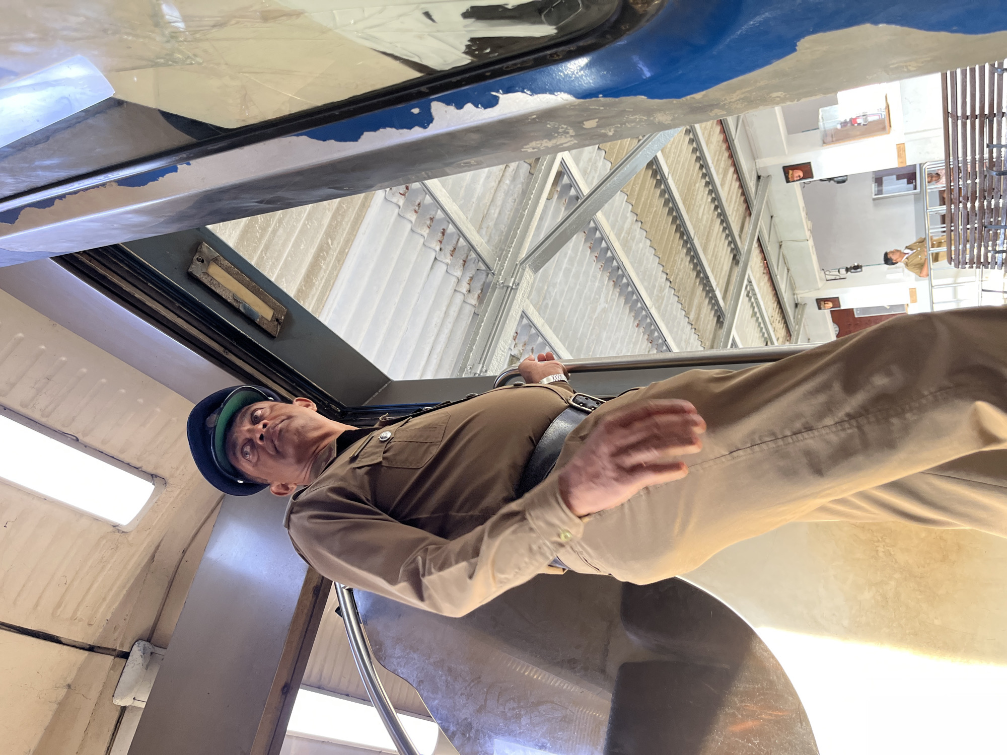 A low-angle, full-body shot captures a middle-aged man in uniform standing inside a train car at a station. He is wearing a khaki uniform with a dark cap, a dark belt, and appears to be looking upwards and to his left. The man's right hand is resting on a railing on the right side of the train car. The train car's interior is visible, with a curved brown partition on the left and the open doorway on the right revealing the train station platform. The station has a corrugated metal roof supported by metal beams. Another person in a similar uniform is visible in the background on the platform, near a bench. The overall scene suggests a moment of observation or transition within a public transport environment. The location is identified as the Ambalangoda railway station in Ambalangoda, Sri Lanka.