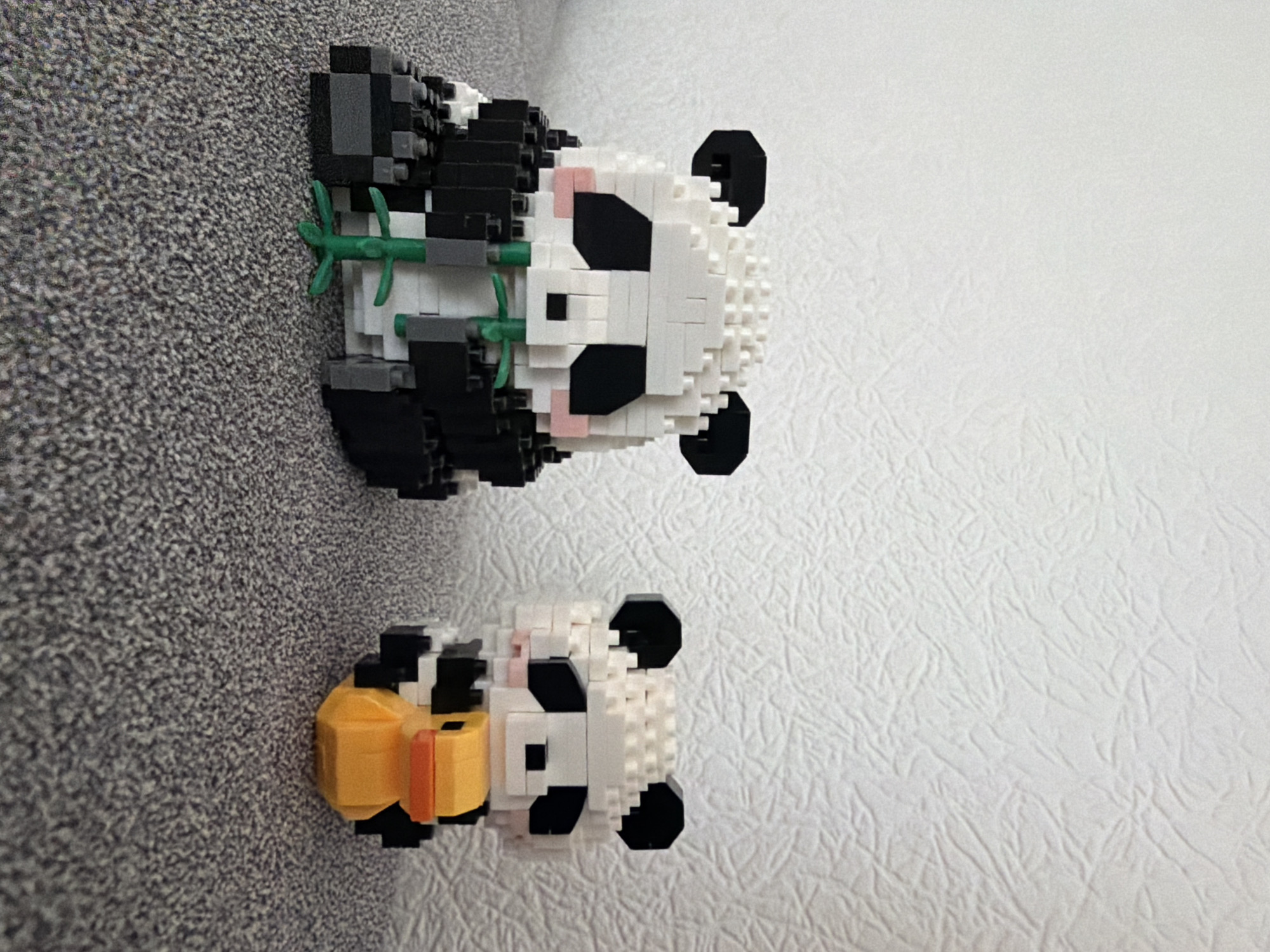 Two panda-shaped figures, constructed from small building blocks, sit on a speckled gray carpet. The larger panda, on the left, is white with black ears, eye patches, and a black torso, and it holds green bamboo stalks in its mouth. A smaller panda, to the right, is identical in construction and color pattern, and it appears to be holding a small orange duck in its mouth. The background is a textured white wall. The image is a static, eye-level shot. There is no indication of the city of Kyiv, Ukraine, in the image.