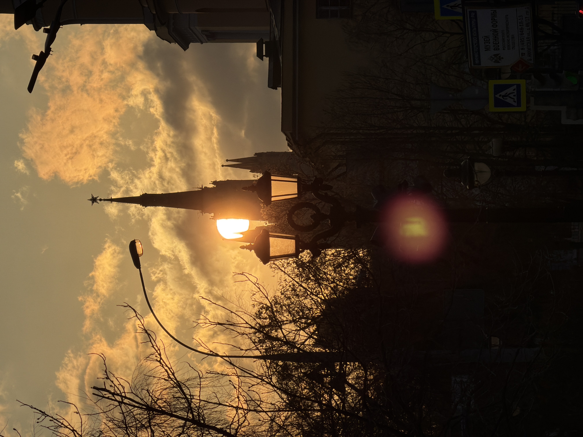 An atmospheric urban landscape captured during sunrise or sunset, characterized by warm, golden-orange light illuminating the sky and silhouetting the city architecture and trees. The scene depicts a dramatic sky filled with textured clouds, glowing with the warm hues of the sun, which is partially obscured by a bright, ornate streetlamp in the midground.
A tall, dark building with a distinctive spired roof and a prominent star on its pinnacle stands silhouetted against the bright sky, reminiscent of Moscow's iconic towers. Other darker buildings line the horizon. In the foreground and midground, the bare branches of trees create intricate, dark patterns against the luminous backdrop. Two streetlights are visible: a modern, curved streetlamp extends from the left towards the sky, and an older, decorative streetlamp shines brightly near the center, casting a strong lens flare.
On the right side of the image, a large information board is visible, along with road signs, including a pedestrian crossing sign. The text on the information board is in Russian and identifies a "МУЗЕЙ ВОЕННОЙ ФОРМЫ" (Museum of Military Uniforms) belonging to the "Российского ВОЕННО-ИСТОРИЧЕСКОГО ОБЩЕСТВА" (Russian Military Historical Society). It also provides a Moscow phone number (8 (495) 648-18-13) and an address: "Б. НИКИТСКАЯ УЛ., д. 46/17, стр.1" (Bolshaya Nikitskaya Street, building 46/17, structure 1). The "МИНИСТЕРСТВО КУЛЬТУРЫ РФ" (Ministry of Culture of the Russian Federation) is also mentioned. The overall mood is serene and dramatic, emphasizing the contrast between the illuminated sky and the darkened urban elements.