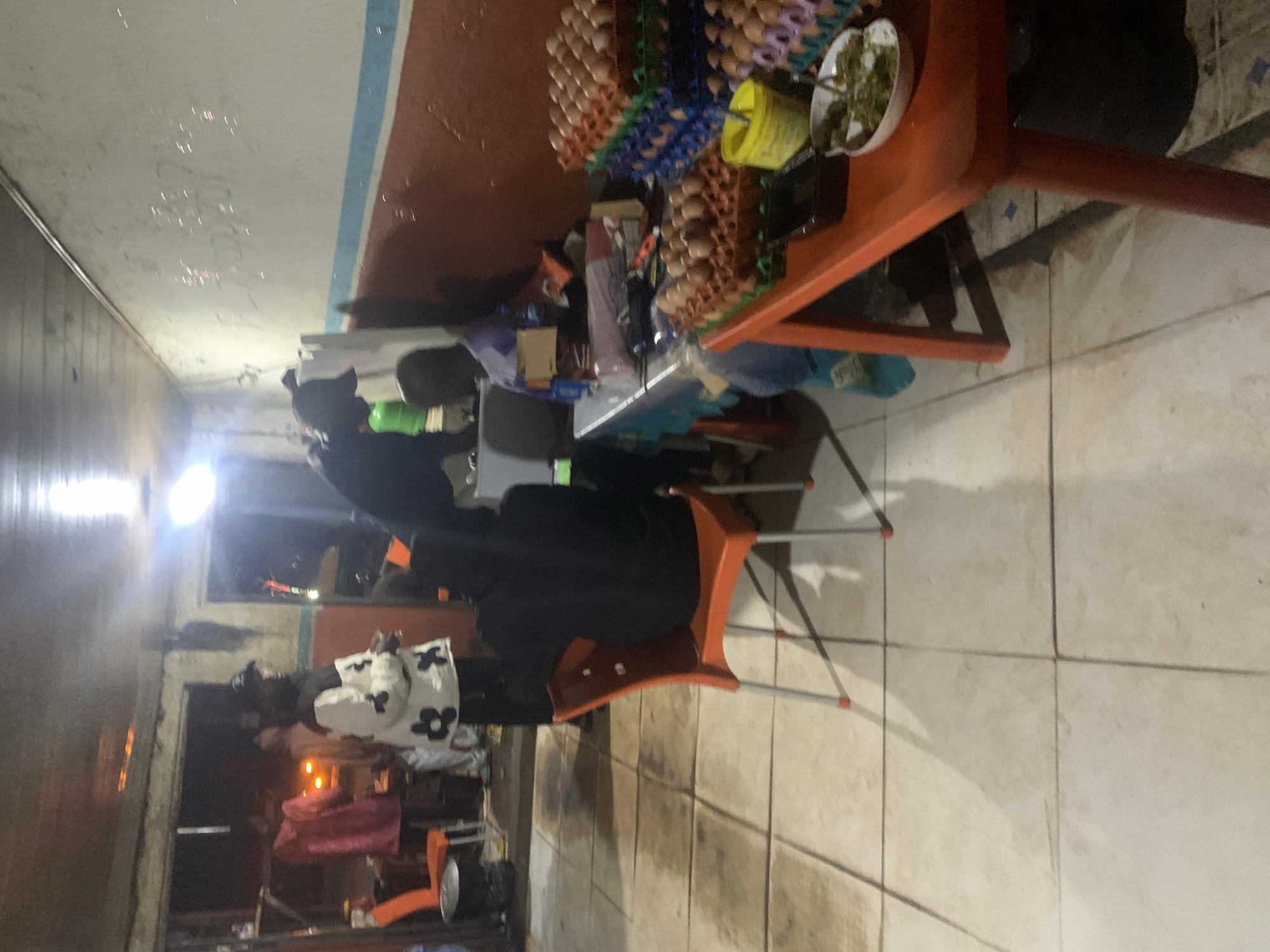 The image depicts a dimly lit indoor scene, possibly a food stall or a small eatery, in Jos, Nigeria. Several individuals are present, some engaged in activities and others seated. In the foreground, a person is seated on an orange plastic chair facing away from the camera. To the left, another orange plastic chair is empty. Further into the scene, a person wearing a white jacket with black floral patterns is visible. Behind them, a person in a pink garment stands near an opening. In the mid-ground, a person wearing a dark hoodie and a baseball cap is leaning over a table. On a table covered with a blue and white checkered cloth, several stacks of eggs in colorful cartons are arranged. To the right of the egg display, a bright orange table is set up, with a small white bowl containing green food items. The walls of the establishment are painted with horizontal bands of red and blue. The floor is tiled and appears wet or stained in places. The overall atmosphere is utilitarian and bustling.