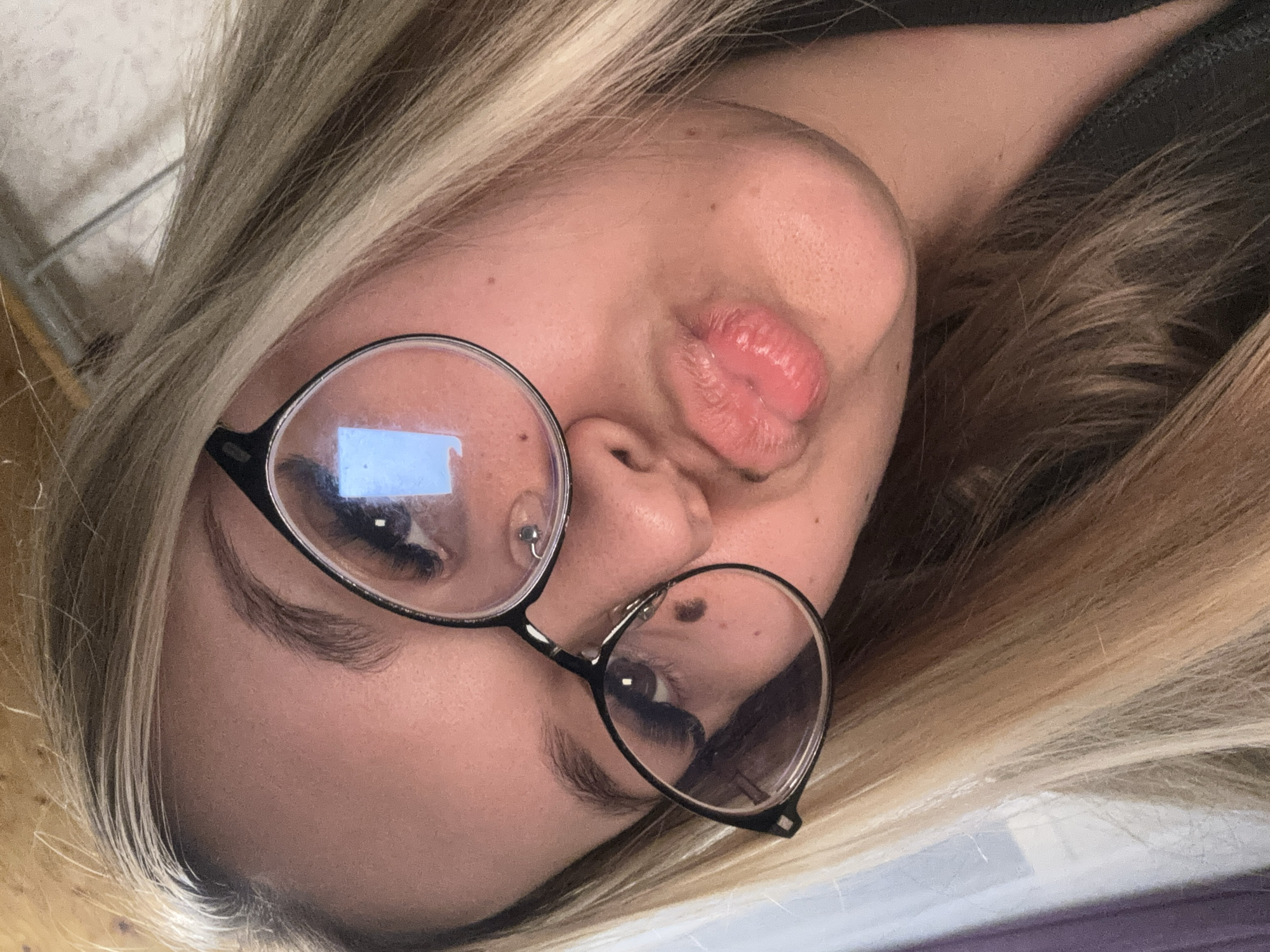 A close-up portrait shows a young woman angled diagonally across the frame, looking towards the viewer. Her head is tilted, with her blonde and light brown highlighted hair flowing around her face. She wears large, round-framed glasses with black rims, through which her eyes are visible. A prominent reflection, appearing like a rectangular light source or screen, is visible in the left lens (viewer's left). Her left ear has a small stud piercing.
The woman's lips are pursed in a playful "kiss" or "duck face" expression, highlighted with a glossy pink product. Her skin appears clear with a few small moles or freckles visible, particularly on her cheeks and forehead. Her eyebrows are well-defined, and she has long eyelashes, suggesting mascara or extensions.
The background is out of focus, showing indistinct light and dark tones, consistent with an indoor setting, possibly a wall or soft furnishing. There are no specific cues in the image to indicate the provided location of Большой Букор, Russia. No visible text is present. The overall impression is a casual, intimate selfie or close-up shot, capturing a moment of playful posing.