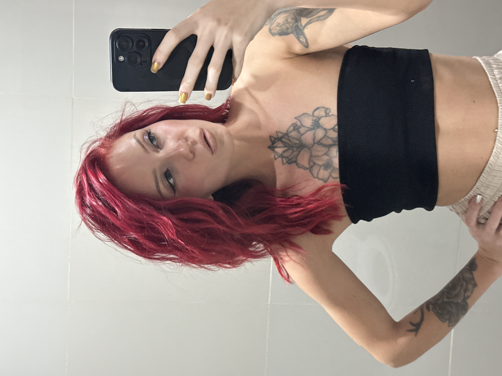 A person with red hair is holding a smartphone in their left hand, taking a selfie. The person is wearing a black bandeau top and beige shorts. Tattoos are visible on both arms and the chest. The background appears to be a white-tiled wall. The location is assumed to be North Kuta, Indonesia.