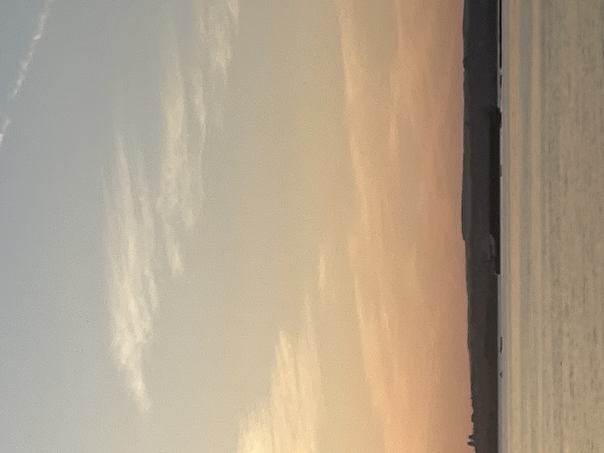 The image depicts a wide, calm body of water, possibly a river or lake, under a soft, gradient sky transitioning from a pale blue at the top to a warm orange and pink near the horizon. Wispy clouds are visible in the upper sky. Along the distant horizon, a dark, silhouetted landmass stretches across the frame, with subtle undulations suggesting hills or a coastline. A long, dark, rectangular object, likely a barge or a docked vessel, is visible on the water near the landmass. Several smaller, dark shapes, potentially other boats or buoys, are also scattered across the water in the distance. The overall scene is tranquil and atmospheric, characteristic of a sunset or sunrise. There are no people or discernible structures beyond the distant landmass and the vessel on the water. The image quality is slightly grainy. The location is identified as Shumeika, Russia.