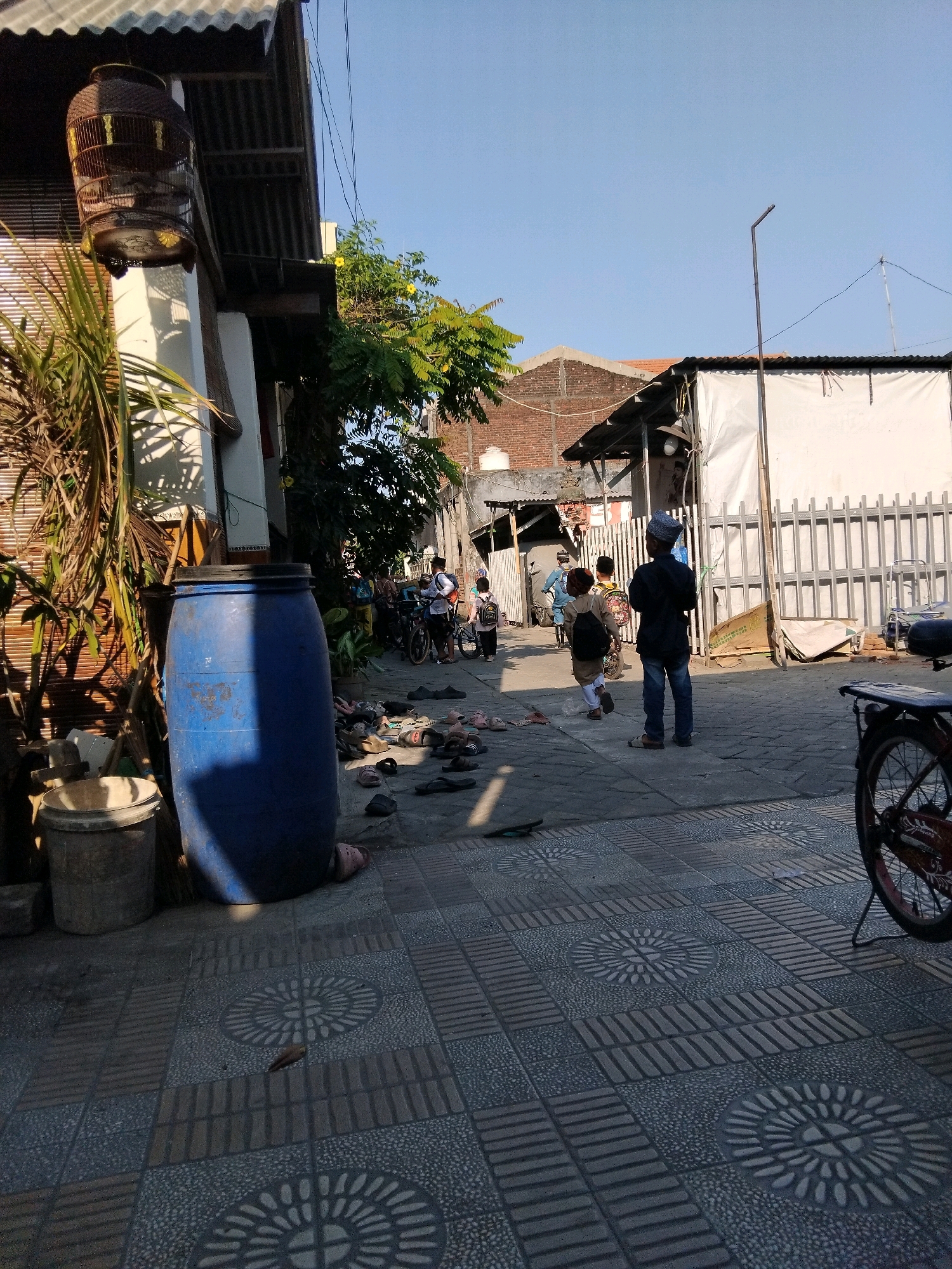 The image depicts a street scene in Kepuh Kiriman, Indonesia, under a clear blue sky. In the foreground, the tiled pavement features circular decorative patterns. To the left, a large blue plastic barrel stands next to a grey bucket and some brooms. A birdcage hangs from the eave of a building with a corrugated metal roof. A palm-like plant is situated near the barrel.
Further down the street, several people, predominantly children, are visible. Some are walking, while others appear to be gathered or about to move. Several pairs of sandals and shoes are scattered on the ground. A bicycle is parked on the right side of the frame. Buildings line both sides of the street, with varying roof materials and construction styles, including brick walls and corrugated metal. The overall impression is a residential street with daily life activities.