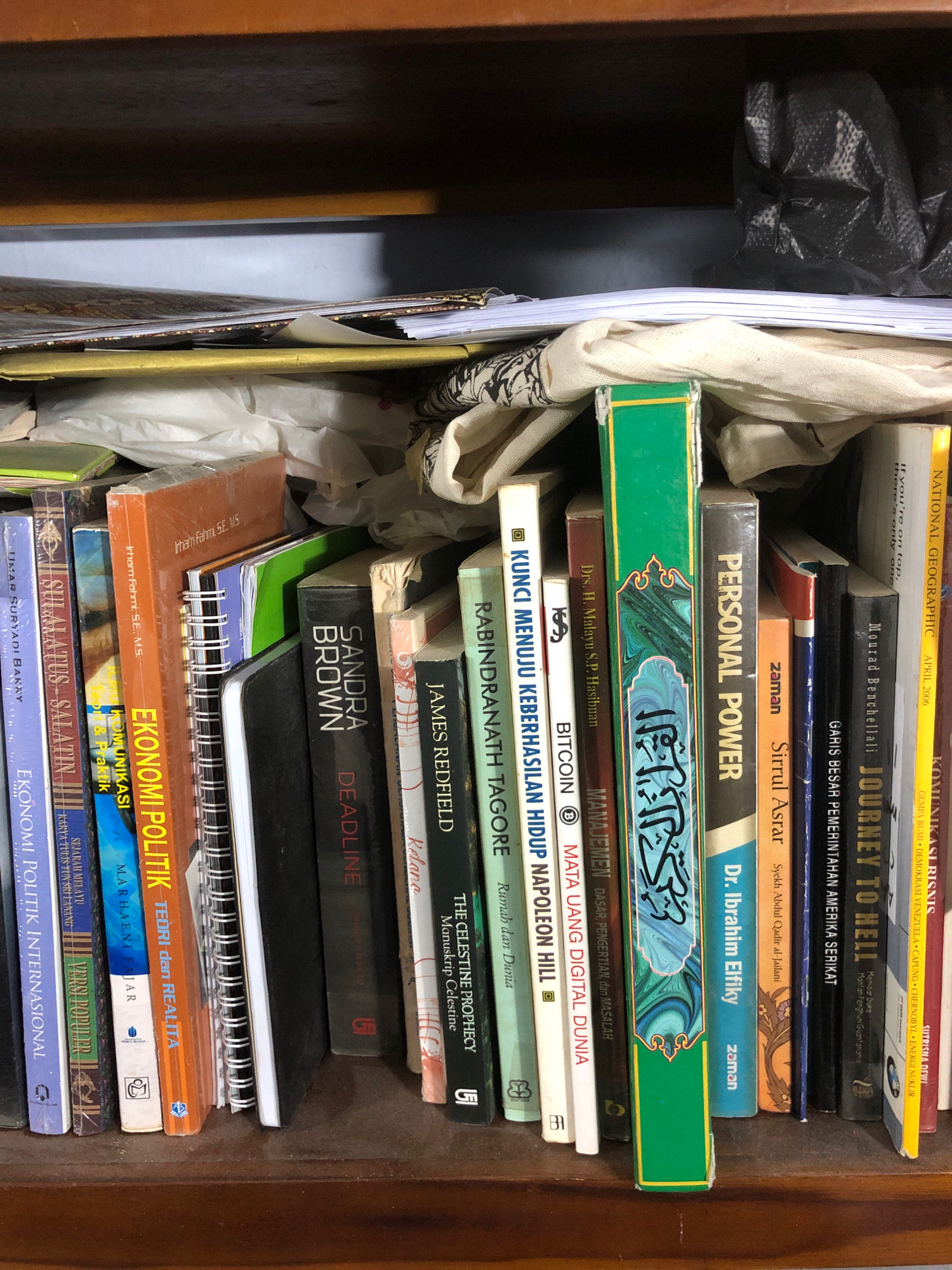 The image shows a close-up of a bookshelf filled with books. The books are of various sizes and colors, and some are stacked horizontally. In the background, there are some crumpled black plastic bags. The shelf appears to be made of wood. The overall impression is one of a well-used personal library. The scene is consistent with a room in Lhokseumawe, Indonesia, given the presence of books in Indonesian and Arabic, and common household shelving.