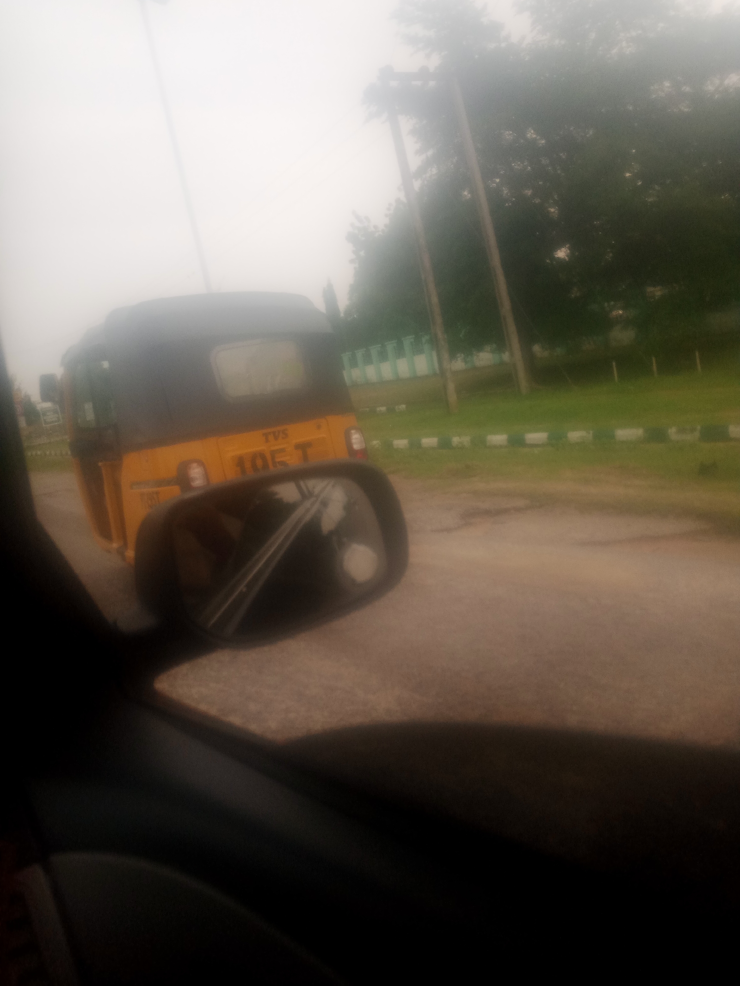 The image captures a side-view of a yellow auto-rickshaw, identified by the "TVS 105 T" marking on its rear, driving on a paved road. The auto-rickshaw appears to be in motion, with its reflection visible in the side-view mirror of the vehicle from which the photo is taken. The environment suggests an outdoor setting with greenery and trees visible in the background. The sky is overcast, contributing to a slightly hazy or foggy atmosphere. The scene is situated in Jalingo, Nigeria, as indicated by the context. The image is taken from inside a vehicle, looking outwards.