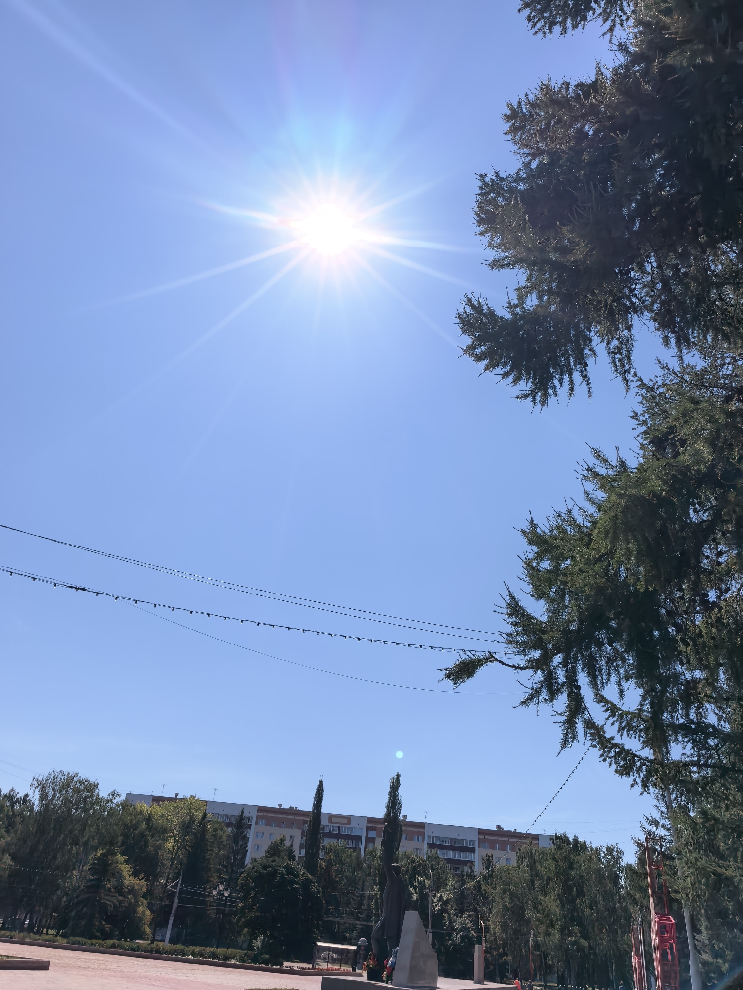 The image shows a bright sunny day in Sterlitamak, Russia. The sky is clear and blue, with the sun shining intensely. In the foreground, there are two large evergreen trees, one on the right and another on the left, their branches extending towards the sky. Between the trees, a string of lights is visible, spanning across the frame. In the mid-ground, a multi-story apartment building with a light-colored facade and darker accents stands behind a dense line of various trees. To the left of the apartment building, a tall, slender tree stands out. Further to the left, in the lower part of the frame, there's a glimpse of a paved area, possibly a park or plaza, with a monument or sculpture visible. The overall scene suggests an urban environment on a clear, warm day.