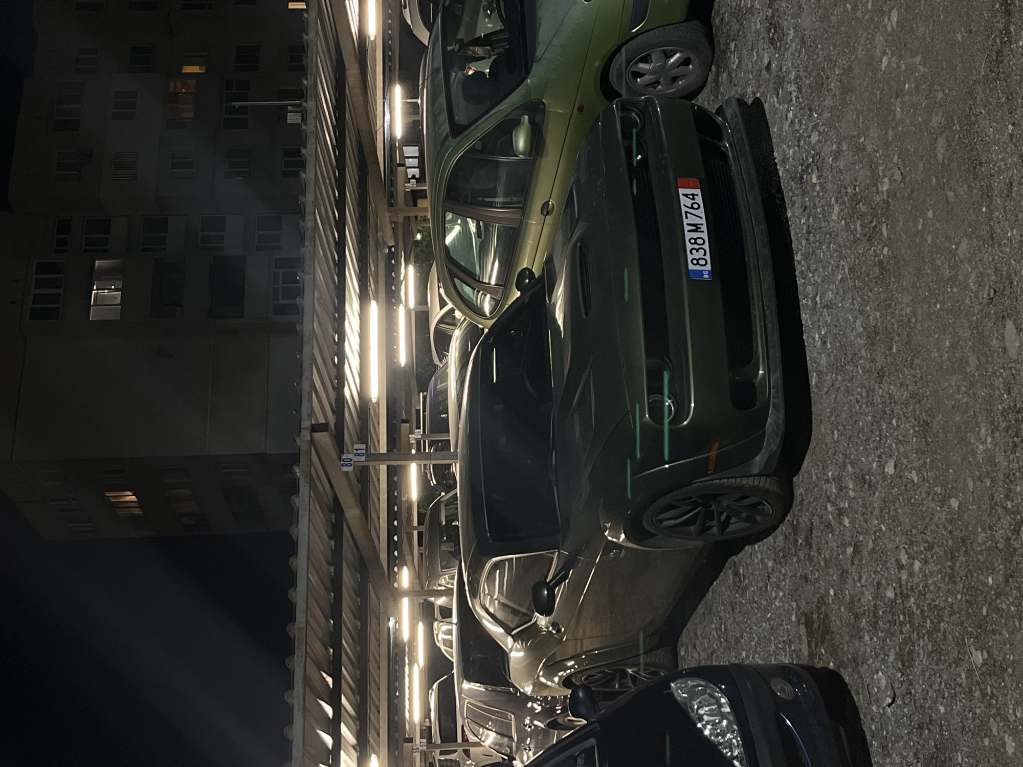 The image depicts a parking garage in Sofia, Bulgaria at night. Cars are parked in a row under fluorescent lighting. The registration plate on a dark-colored car in the foreground reads "838 M 764," which identifies it as a Bulgarian vehicle. A building is visible in the background, lit by a few windows. The ground outside the garage appears to be paved with asphalt.