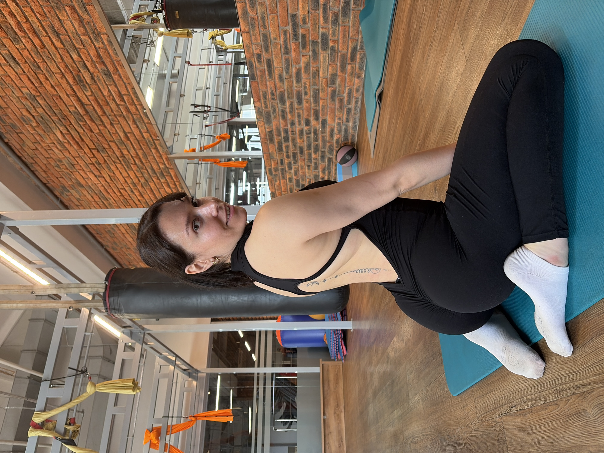 The image shows a woman in a black jumpsuit and white socks sitting on a teal yoga mat in a gym. The gym has a wooden floor, brick walls, and aerial yoga equipment hanging from the ceiling. A punching bag is visible in the background. The woman is facing away from the camera and looking over her shoulder with a smile. Her back has a tattoo that runs vertically down her spine. The scene is located in a gym in Chaikovsky, Russia.