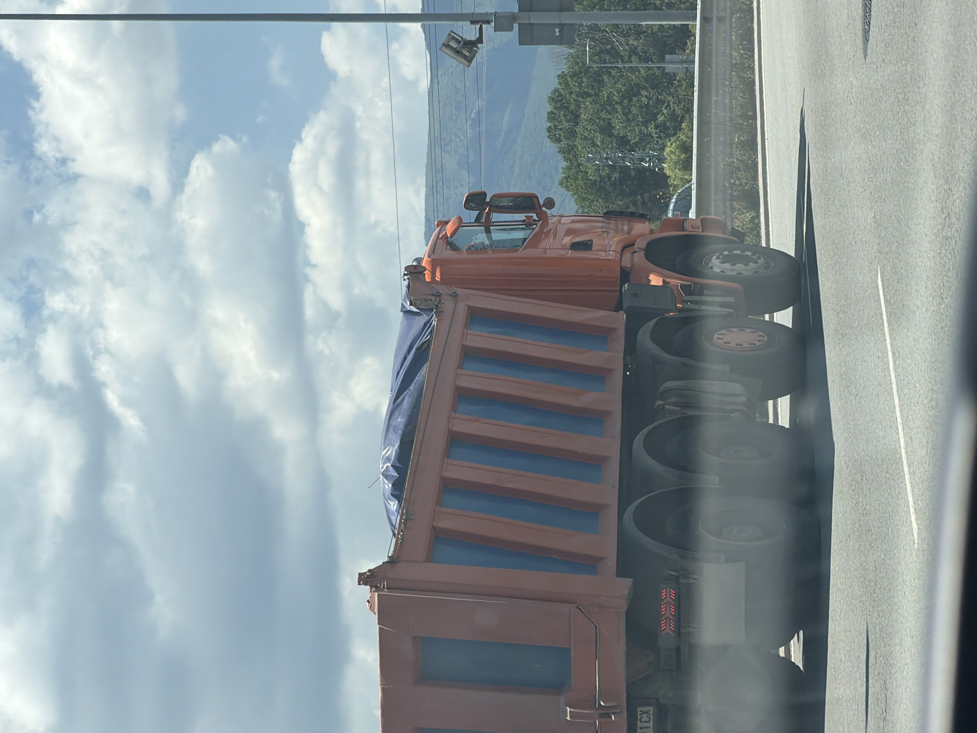 An orange dump truck is driving on a highway in Sofia, Bulgaria. The truck has a blue tarp covering its cargo bed, and it is emitting a plume of white smoke from its rear wheels. The truck's license plate is visible, reading "1 CX". The highway is paved with asphalt and has white lane markings. In the background, there are green trees and mountains. The sky is cloudy with patches of blue sky. There is a lamppost with a security camera attached to it on the right side of the image.