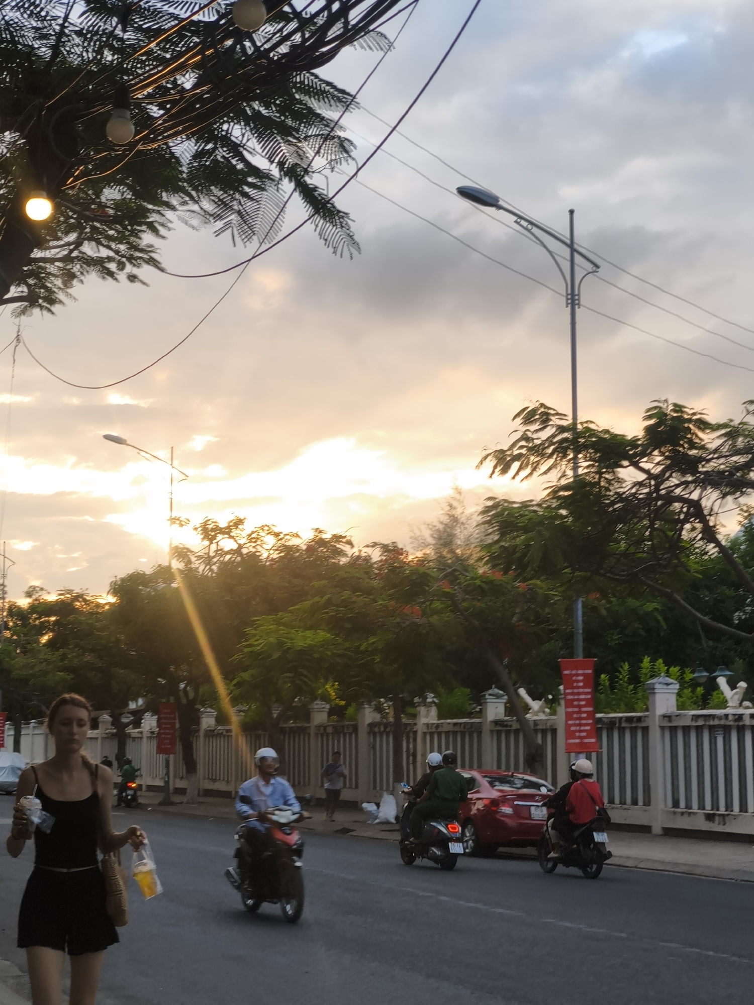 This is a street scene in Nha Trang, Vietnam, during sunset. A young woman in a black tank top and shorts walks towards the viewer, carrying a drink and shopping bags. Behind her, several motorbikes are on the road. One motorbike has a rider in a blue shirt and white helmet, while another has a rider in a green shirt and helmet. A red car is parked on the side of the road. The sky is cloudy with the sun setting, casting a warm glow over the scene. Trees line the street, and a streetlight is visible in the background. The street appears to be paved, and there is a sidewalk with a fence along the right side of the road.