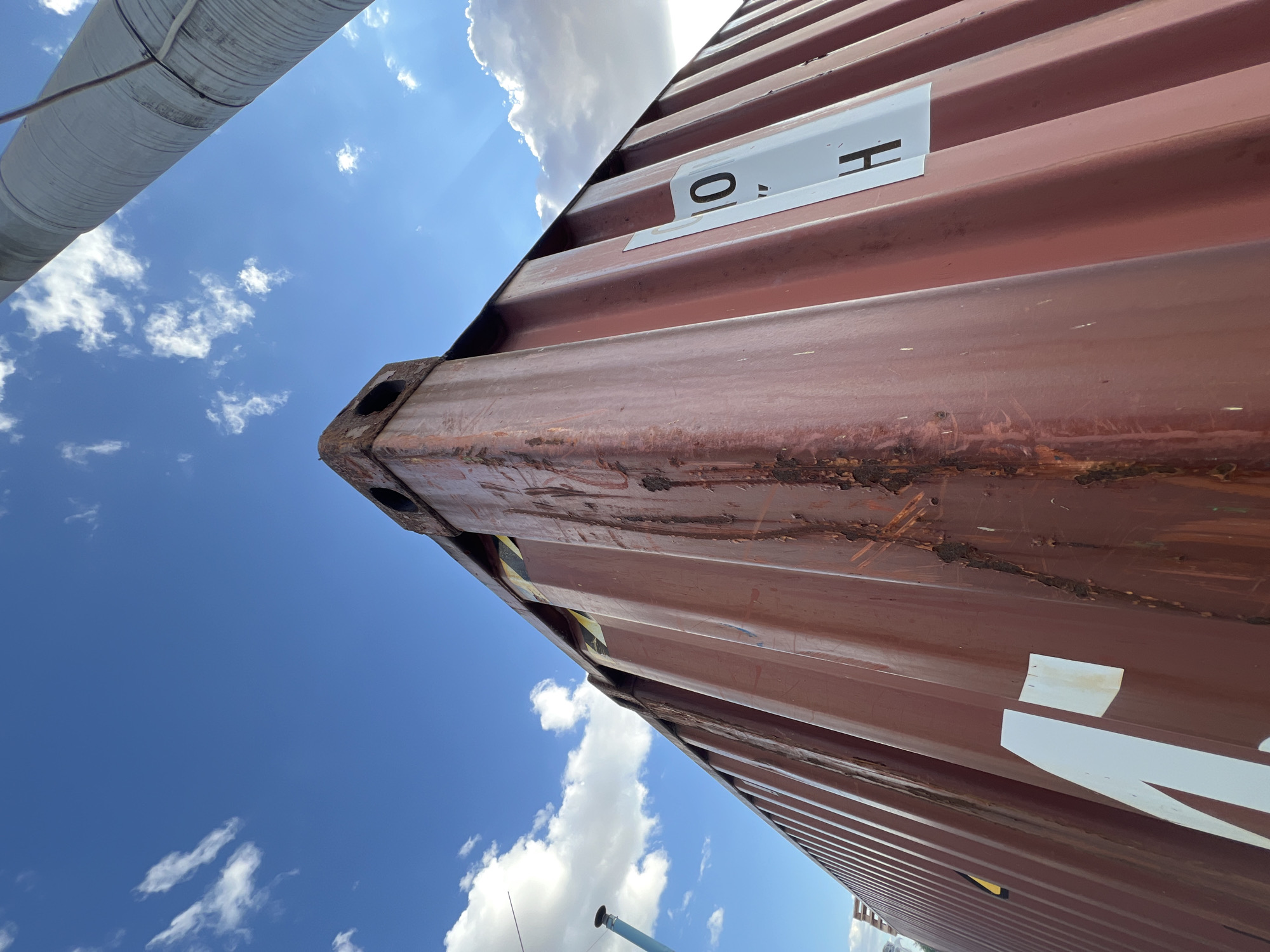 This image captures a close-up, low-angle view of a large, rusty, corrugated metal shipping container against a bright blue sky dotted with fluffy white clouds. The container is a deep maroon or brown color, showing signs of wear and tear with visible rust streaks and some peeling paint. A white label with black stenciled letters "O H" is affixed to the side of the container. Another larger white marking, partially visible, appears to be part of a word or logo.
The perspective is from below, looking upwards, emphasizing the massive scale of the container. Part of a large, ribbed, light-colored cylindrical structure, possibly a pipe or a silo, extends into the frame from the top left, also angled against the sky. The bright sunlight suggests it is daytime, likely mid-morning or afternoon, and the clear sky with scattered clouds indicates fair weather.
The scene appears to be an outdoor industrial or port area, given the presence of the shipping container. There are no people visible. The overall impression is one of industrial infrastructure under a clear sky. The location context provided is Újezd u Brna, Czechia, which might suggest a logistics hub or a similar facility in that region.