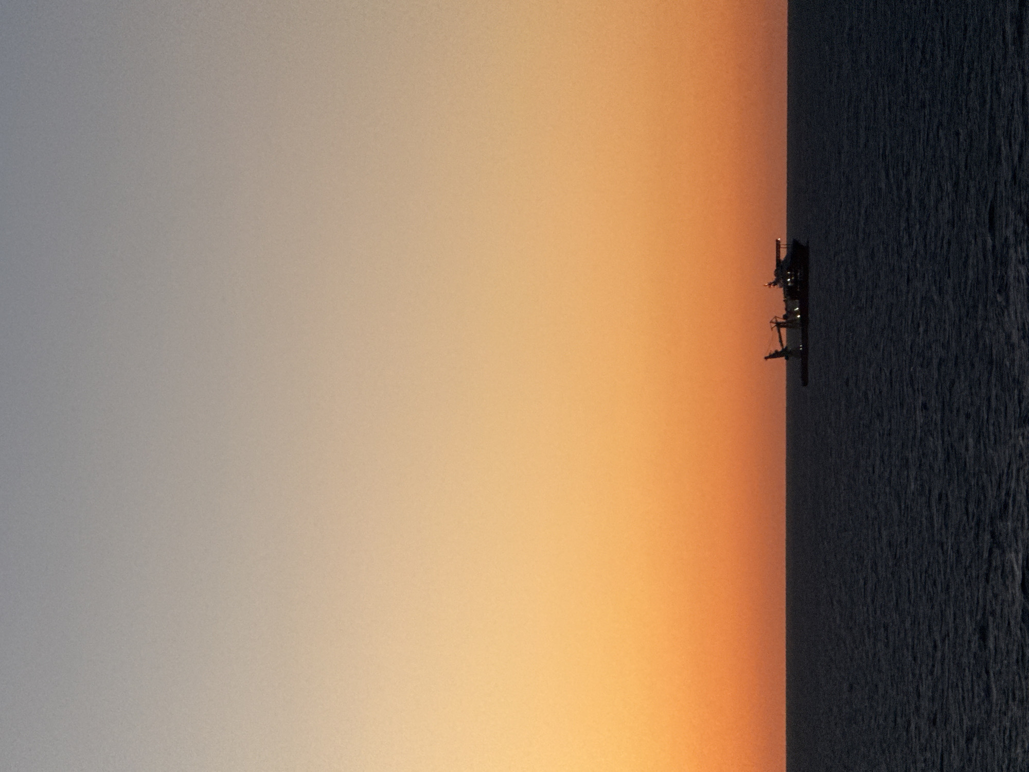 The image depicts a body of water on the right side, transitioning to a gradient of orange and grey towards the left. A structure is visible on the horizon, positioned between the water and the orange sky.