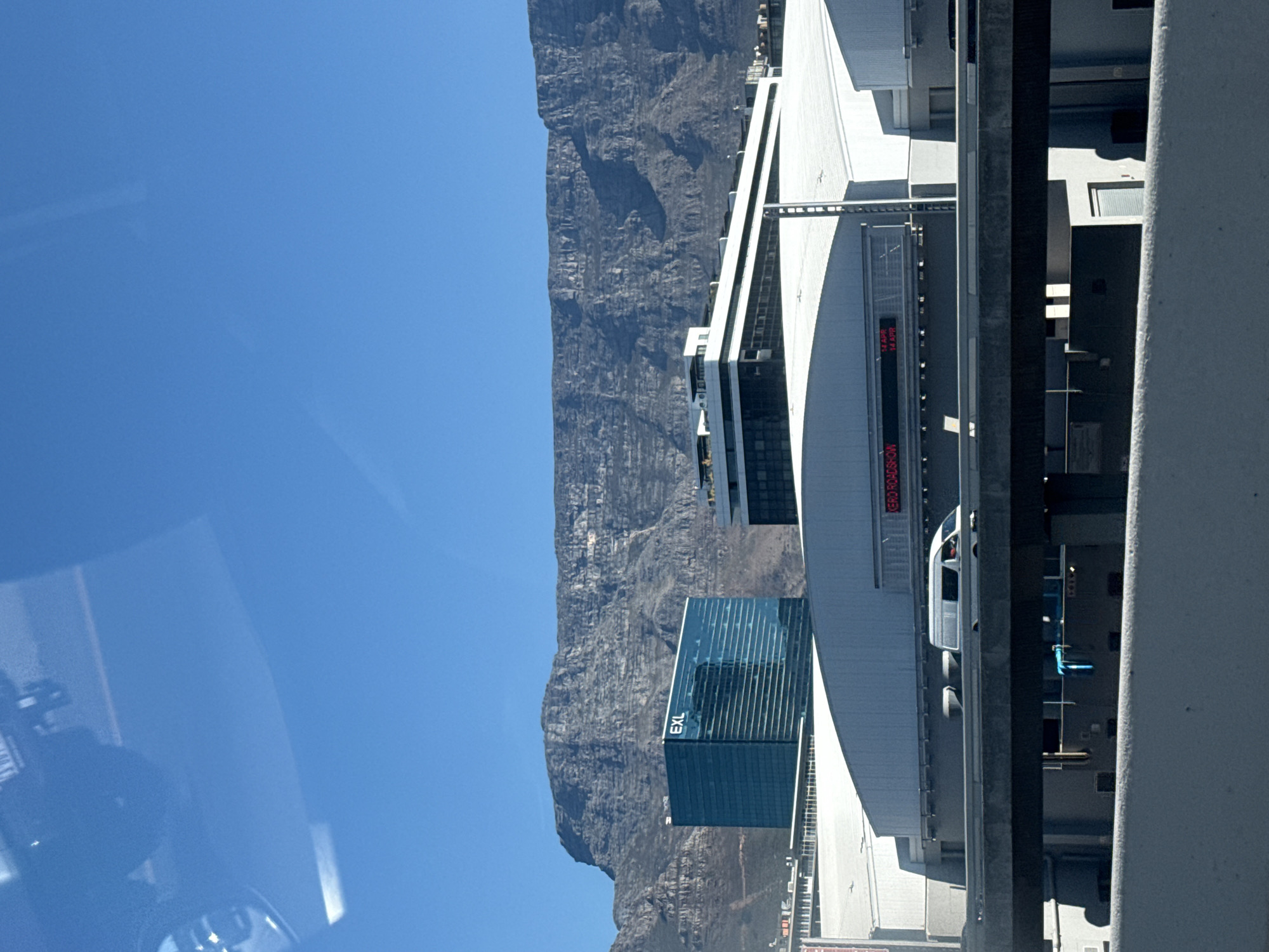 The image depicts a view of Cape Town, South Africa. In the background, Table Mountain dominates the skyline under a clear blue sky. In the foreground, modern architecture is visible, including a tall glass building with the letters "EXL" displayed vertically. A white structure with a digital display showing the word "ROADSHOW" is positioned next to a road. The viewpoint appears to be from inside a vehicle, as evidenced by a partial reflection on the left side of the image.