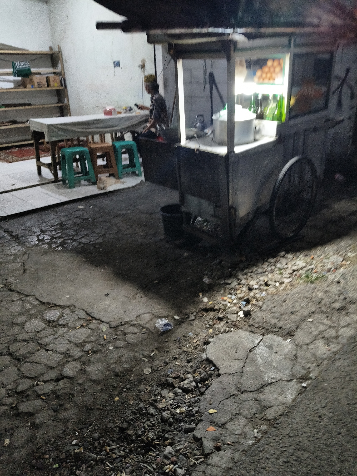 The image depicts a nighttime street scene in South Tangerang, Indonesia, likely in a residential or commercial area with a mixture of paving and cracked asphalt. A street food cart is prominently featured, illuminated by interior lighting, showcasing bottles of drinks and a pot. A person wearing a peci (a cap common in Indonesia) is seated, appearing to be a vendor or customer. Nearby, a table is set with chairs and a tablecloth, suggesting a dining area. A light-colored cat is visible resting on the ground near the chairs. The environment is somewhat cluttered, with debris on the ground and shelves with items in the background. The lighting is low, contributing to a somewhat grainy image quality.