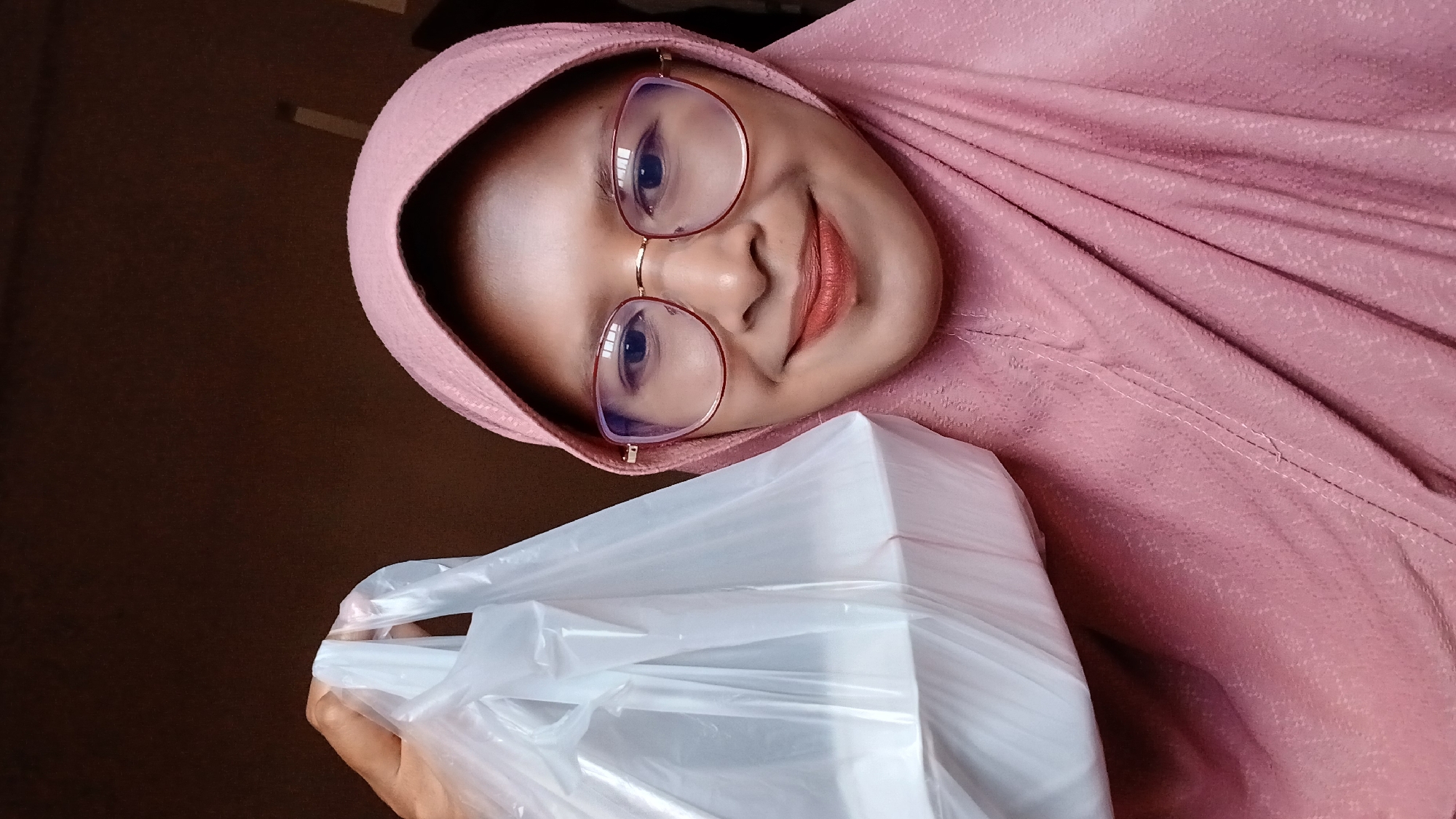 A woman wearing a pink hijab and glasses smiles at the camera while holding a white plastic bag. The background is a dark, solid color. The location is suggested to be Lhokseumawe, Indonesia, but this is not discernible from the image itself.