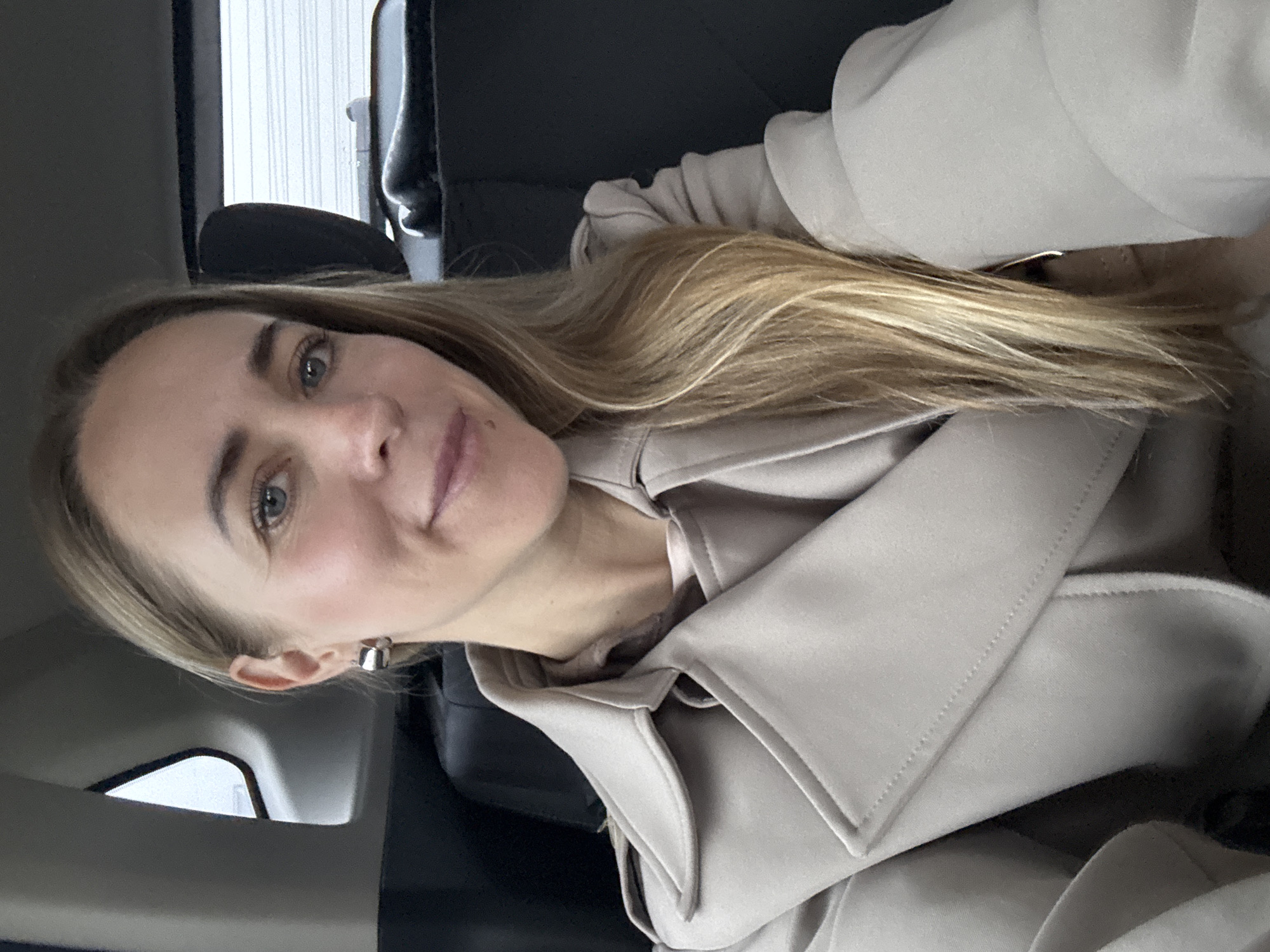 A close-up portrait, likely a selfie, captures a woman seated inside a vehicle. The woman has long, flowing blonde hair, light blue eyes, and a fair complexion. She is looking directly at the camera with a subtle, pleasant smile. She is wearing a light beige or cream-colored coat or jacket, possibly a trench coat, with visible lapels and structured fabric. A silver-toned hoop earring is seen on her left ear. The background is softly blurred, showing the dark interior of a car, including what appears to be a car seat or headrest, and a window to the left, which reveals an indistinct, bright outdoor view, suggesting daytime. The overall impression is calm. No text is visible in the image. This photo is provided with the location context of Chaikovsky, Russia.