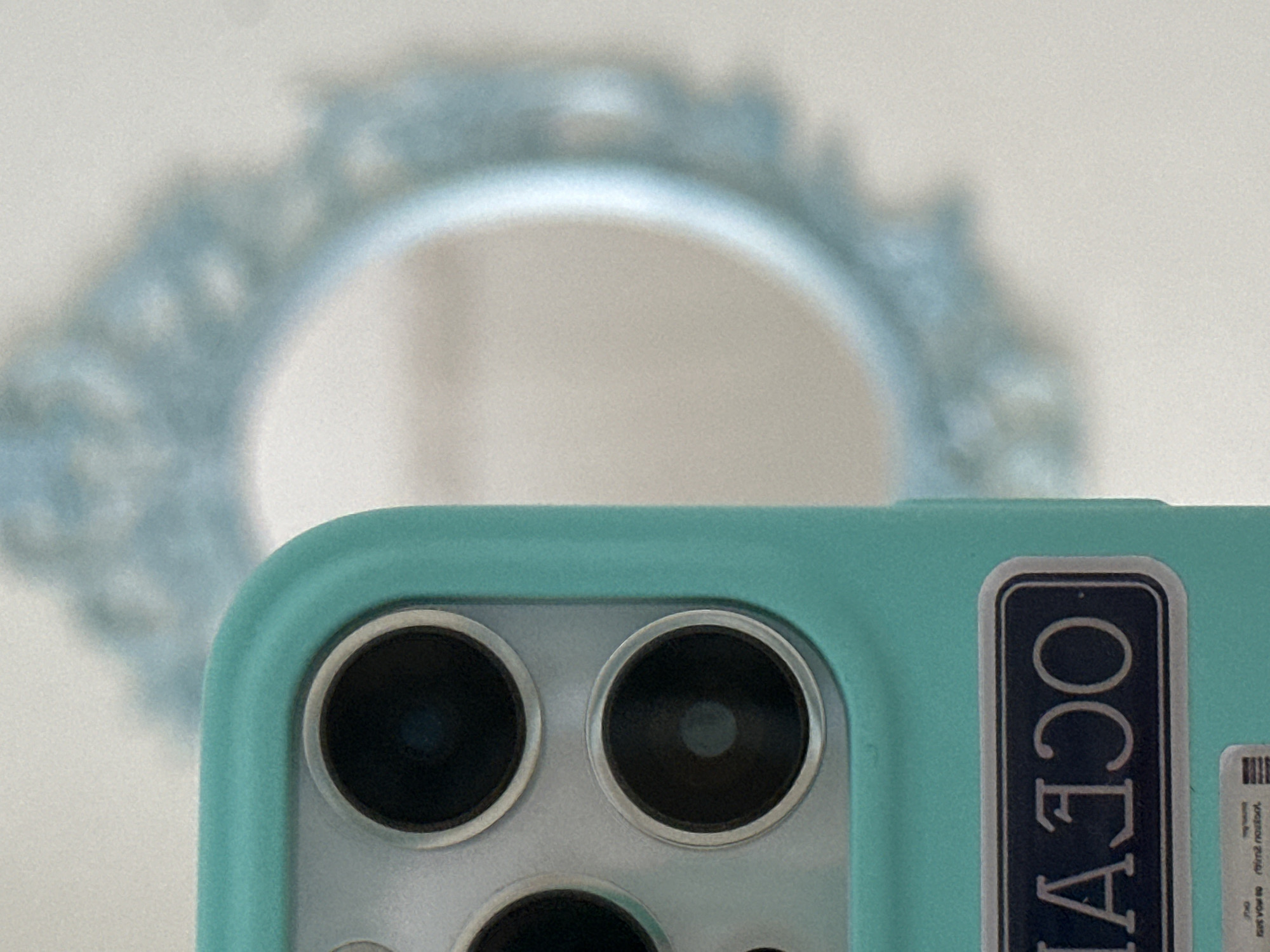 The image shows a close-up of a light teal phone case covering a phone camera with two visible lenses. A sticker with the word "OCEAN" is attached to the case. In the background, there's a blurred object, possibly a decorative mirror. The image doesn't allow for the determination of the location in Saint Petersburg, Russia, or any details about people, actions, or interactions.