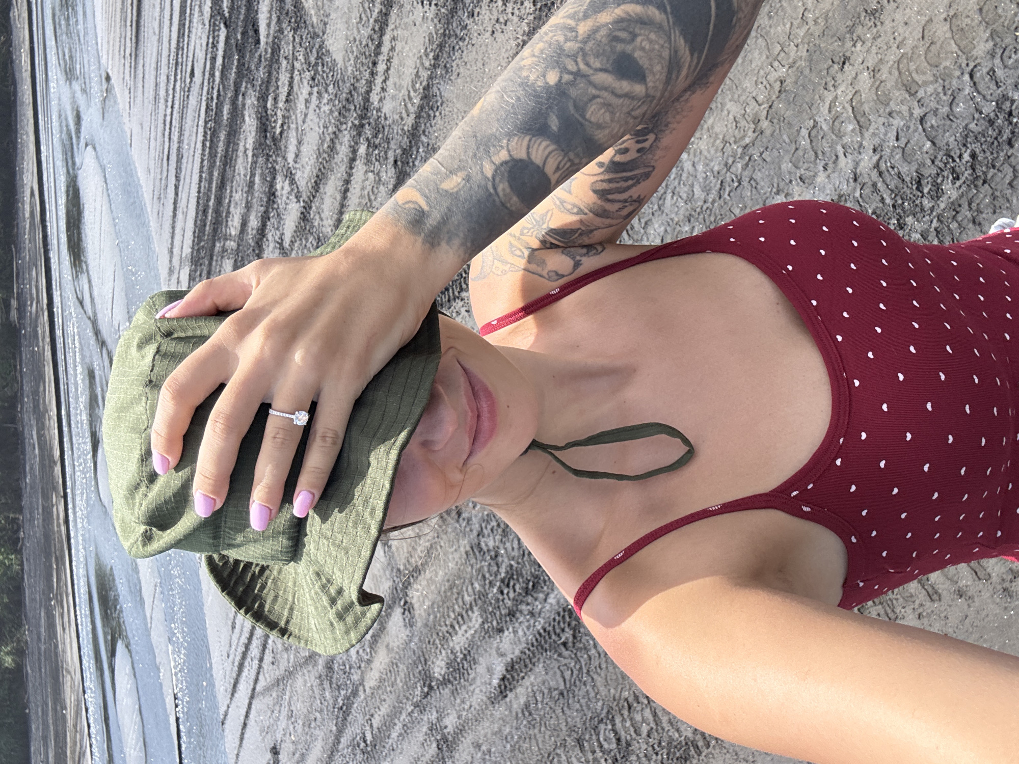 The image depicts a woman with light skin, likely in a selfie, outdoors. She wears a maroon swimsuit with white polka dots and a green bucket hat, partially covering her face. Her left arm, visible in the upper left, is heavily tattooed. She is wearing a ring on the ring finger of her left hand, which is resting on the hat. The background shows a textured gray surface, possibly concrete. The location is Kerambitan, Indonesia, based on the available information. The lighting is bright, suggesting daylight.