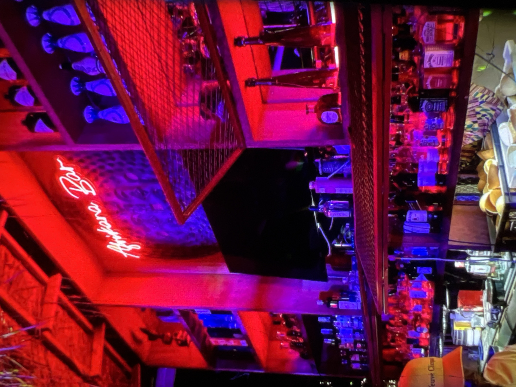 The image depicts the interior of a dimly lit bar, illuminated by vibrant red and purple neon lights. A neon sign reading "Afrikana Bar" in white script is prominently displayed on the wall. Shelves are stocked with numerous bottles of alcohol, including recognizable brands like Jack Daniel's and Gentleman's. A flat-screen television is mounted on the wall behind the bar. In the foreground, part of a person's head wearing an orange hat with the text "Veuve Clicquot" is visible, suggesting they are likely a staff member or patron. The overall ambiance suggests a lively nightlife setting, possibly in Lagos, Nigeria, known for its vibrant bar scene.