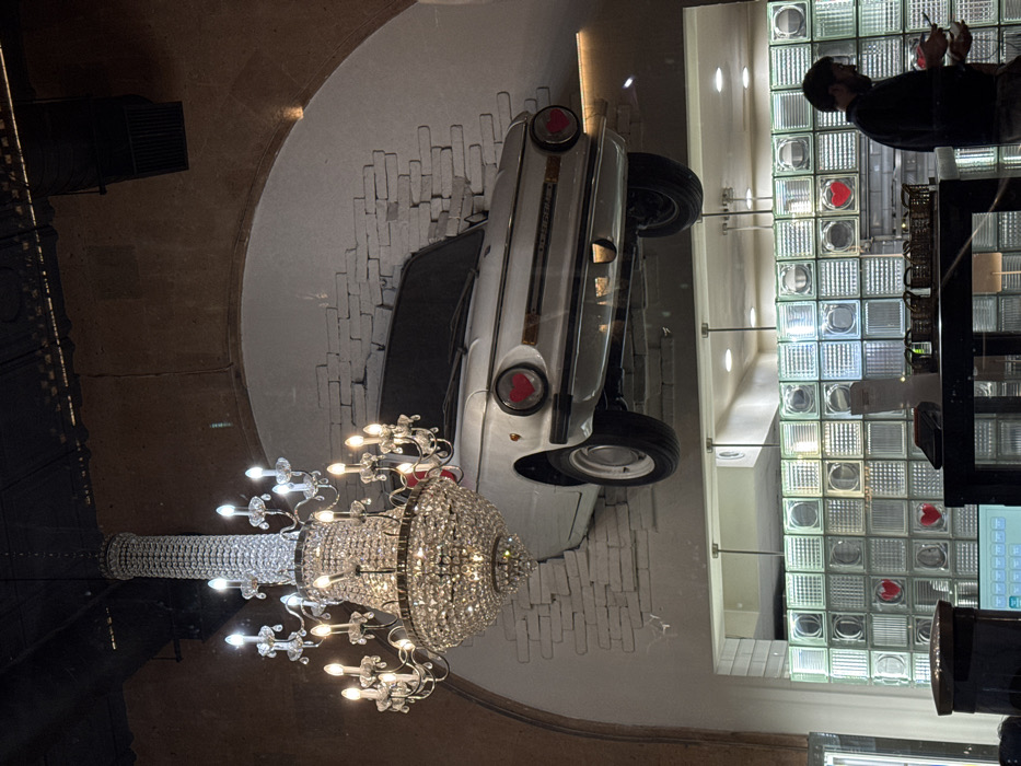 The image displays an interior view of a commercial establishment in Yerevan, Armenia. A large, multi-tiered crystal chandelier with numerous illuminated bulbs hangs from a dark ceiling in the upper left portion of the frame. Directly below and to the right of the chandelier, a white compact car is mounted horizontally on a white-painted brick wall within a large archway, creating the illusion of partial penetration. Red heart symbols are affixed to the car where the headlights would typically be. Below this car installation, a counter is visible, featuring multiple small baskets and a digital screen. Behind the counter, a wall composed of translucent glass blocks is present, with several individual blocks containing red heart symbols. A male individual with dark hair and a beard is partially visible on the right, positioned behind the counter and holding a handheld device.