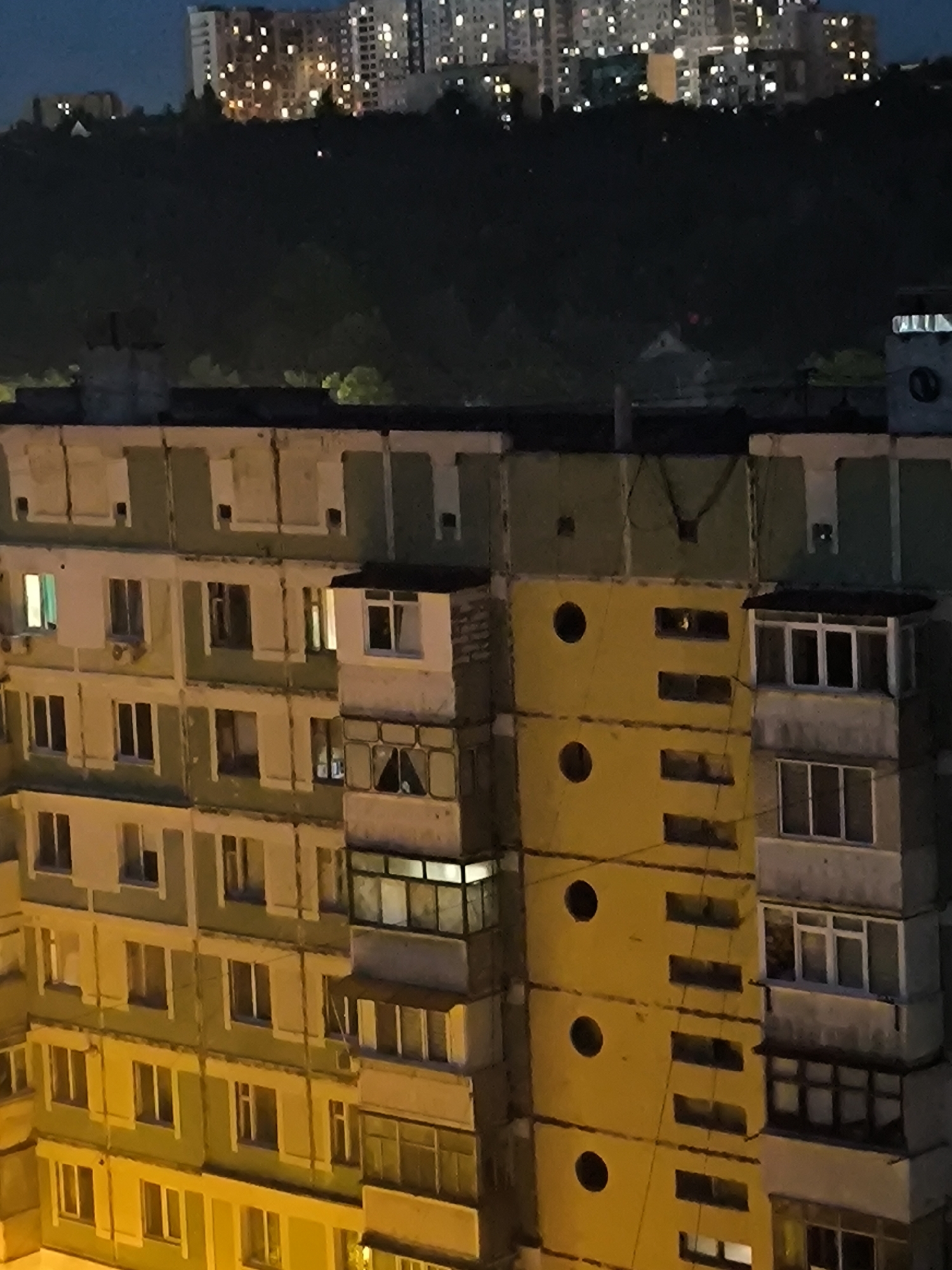 The image shows a nighttime view of apartment buildings in Chișinău, Moldova. The buildings are multi-story and appear to be of Soviet-era construction, with a distinctive yellow and green facade. Many windows are lit, suggesting the presence of residents. In the background, more distant buildings with illuminated windows are visible on a hillside. The overall scene conveys an urban residential environment at night, with artificial lighting from the buildings illuminating the foreground and casting a glow on the surrounding darkness.