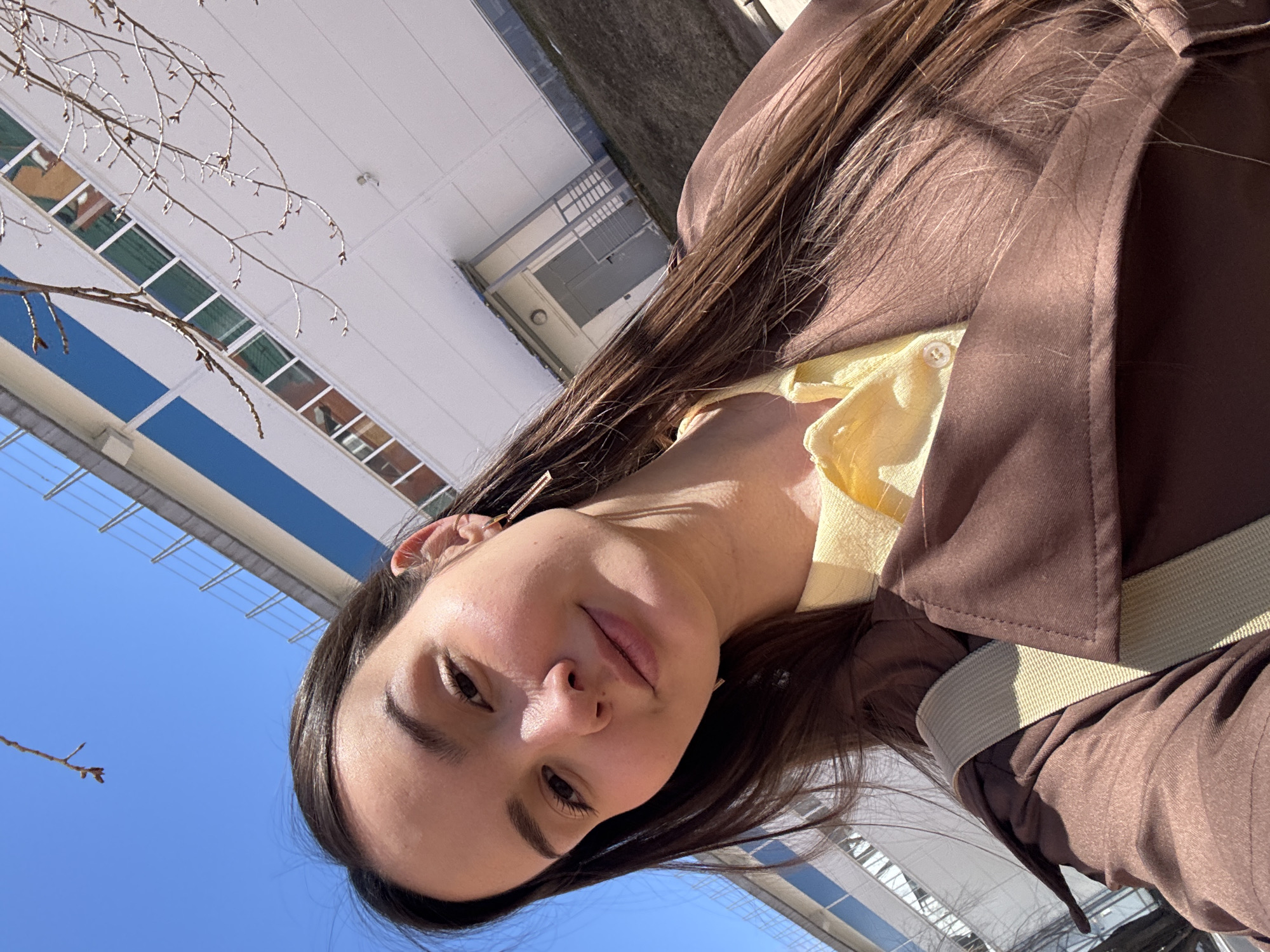 A low-angle, outdoor selfie photograph shows a brunette woman with an off-white dress, brown jacket, and tan belt, facing the camera with the sky and a multi-story building with blue and white facades in the background. The building has multiple rectangular windows. The sky is clear blue. A bare tree branch appears in the lower left corner. The location is Chaikovsky, Russia.