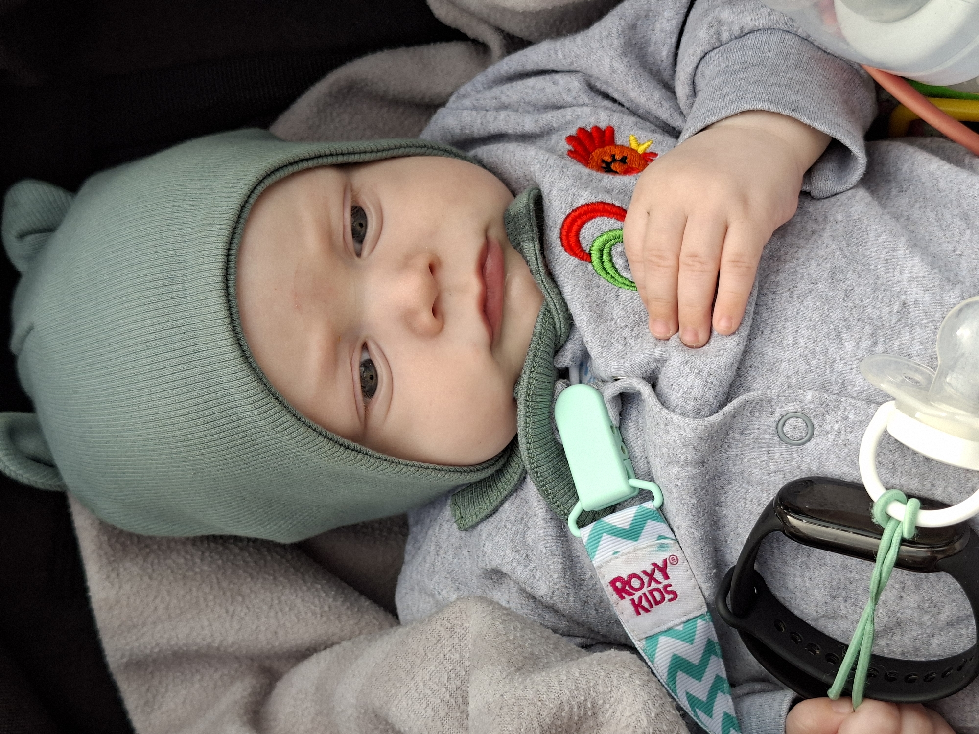 A baby wearing a light green bonnet with small ears lies on a light gray blanket inside a stroller. The baby is dressed in a gray long-sleeved onesie with an embroidered design of two circles and a crab near the chest. The baby's hand rests on the onesie. A teal pacifier clip with a white strap patterned with teal chevrons is attached to the onesie. The strap has the text "ROXY KIDS" printed in pink. A black fitness tracker is tied to a white pacifier ring with a light green cord. The baby is looking up with open eyes. The stroller's interior is black.