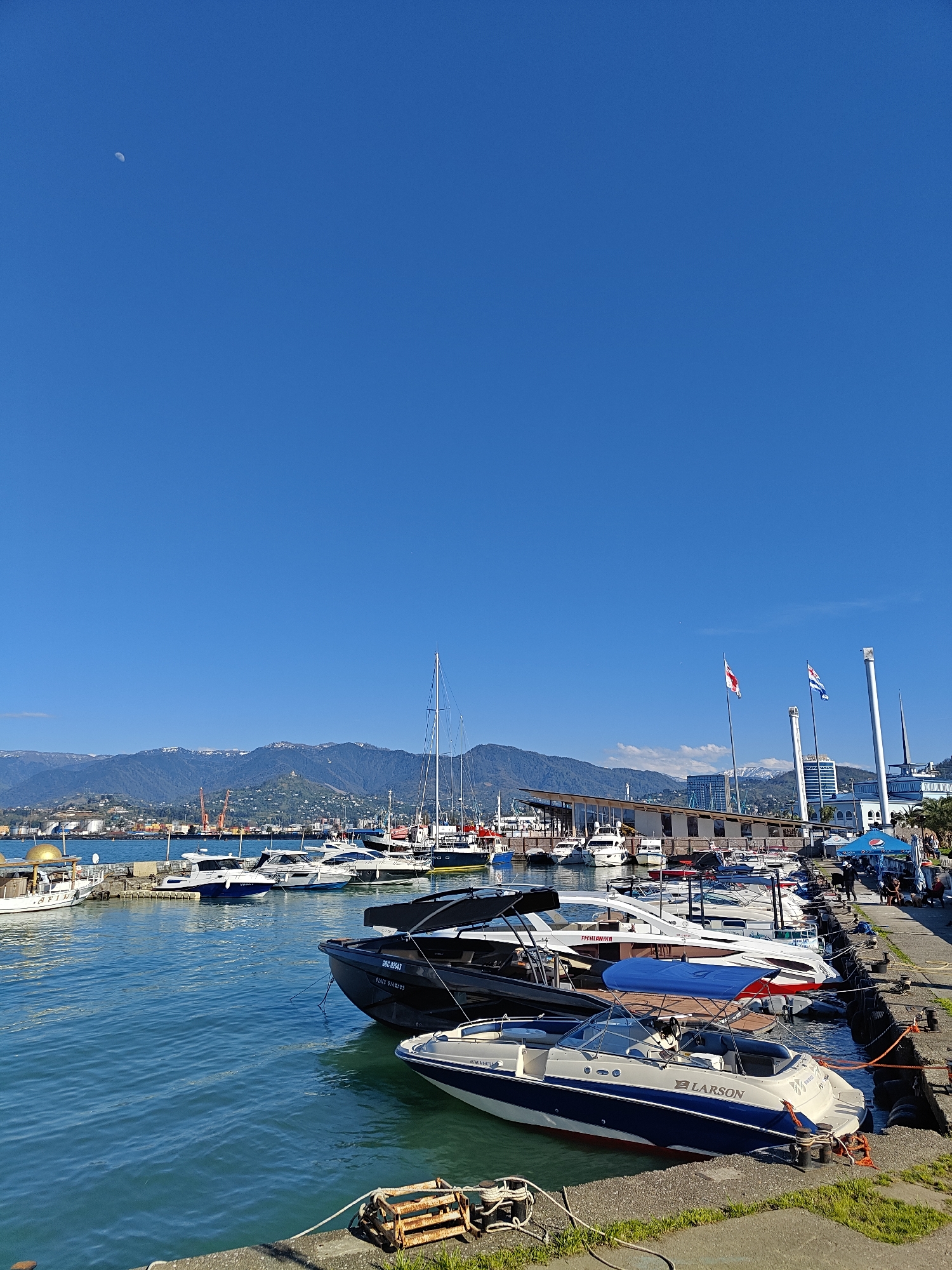 The image depicts a waterfront scene with several boats docked along a concrete pier in Batumi, Georgia. The foreground features a Larson boat in the lower right corner, alongside other various docked boats. The water is a clear turquoise color.
In the background, there are buildings, mountains with some visible snow on their peaks, and structures of the Batumi Sea Port. The clear blue sky occupies most of the upper portion of the image, with a crescent moon visible.
People are present along the pier but are not a central focus. Flags are waving in the air behind the marina buildings.